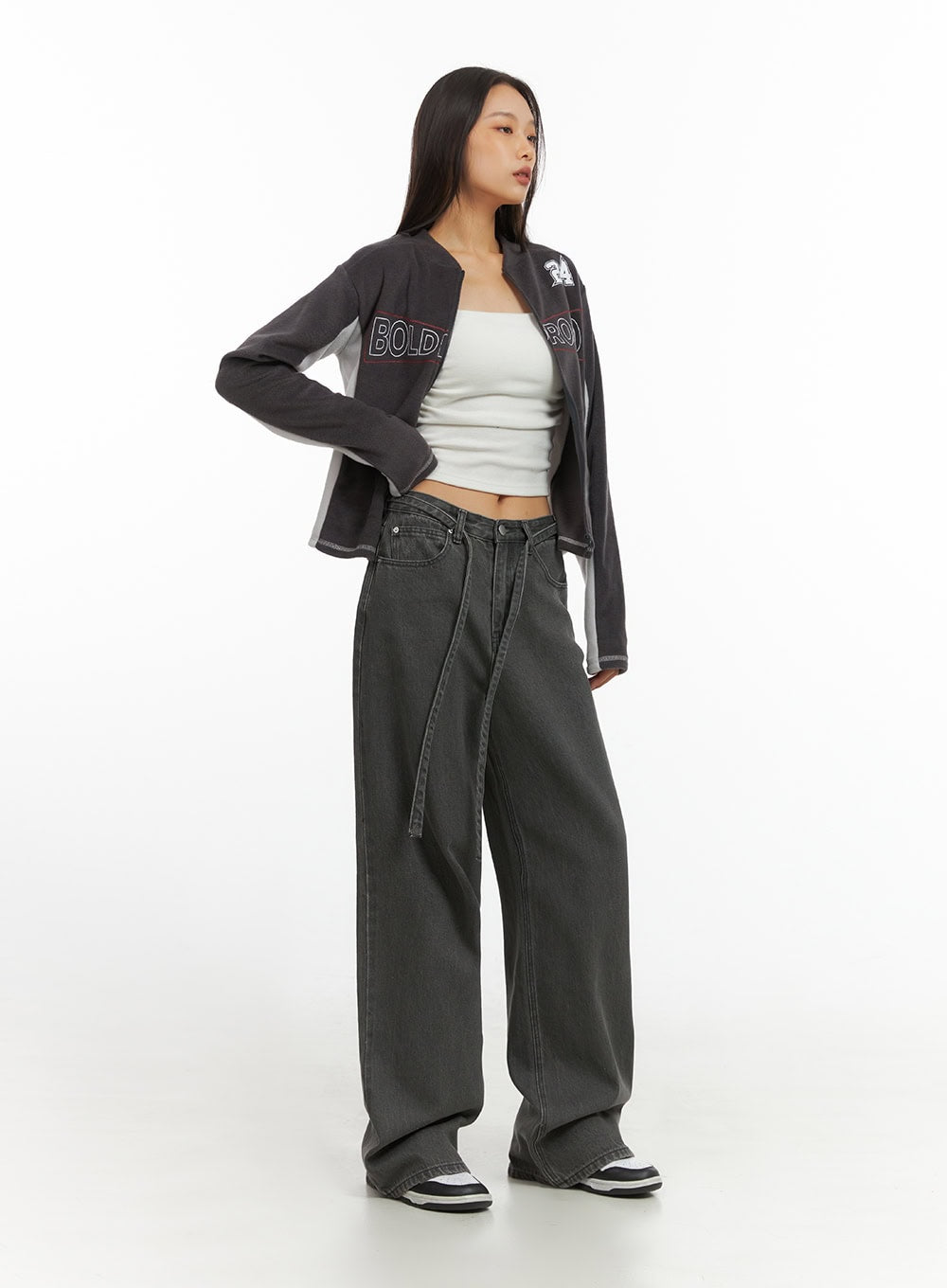 Emery Cotton Strap Baggy Jeans IO409 in dark gray, three-quarter front view on model, baggy straight-fit denim with strap, styled with open varsity jacket and white tube top.