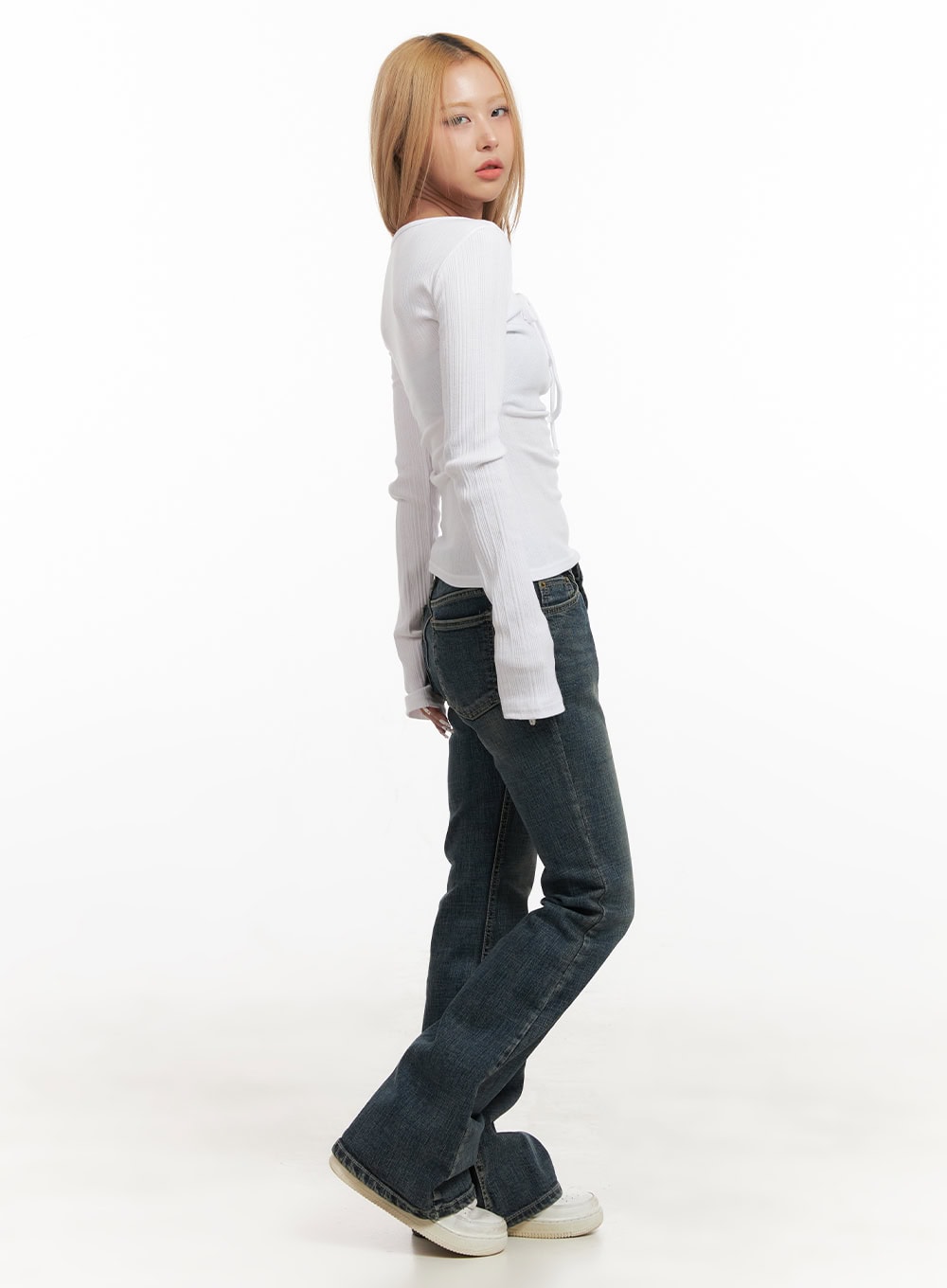 Natalia Low-Rise Flared Jeans CJ508 in dark blue, side profile view on model looking back, washed bootcut denim showing slim thigh to flared leg, paired with white ribbed long-sleeve top and sneakers.