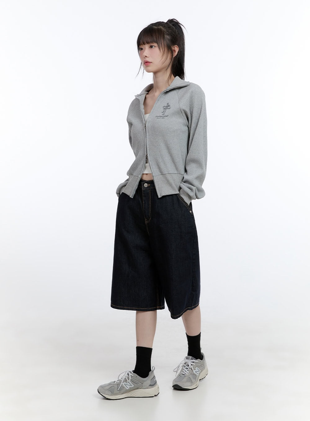 Dora Stitched Baggy Jorts CG413 in dark blue, three-quarter front angle on model looking aside, wide-fit denim jorts with contrast stitching, layered under grey zip jacket.