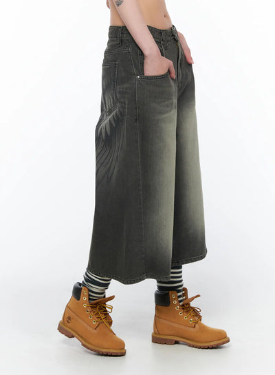 Vintage Style Baggy Denim Shorts CA524 in dark gray, side view on model, washed cotton wide-leg silhouette with wing graphic on thigh, hand in pocket, paired with striped socks and tan boots.