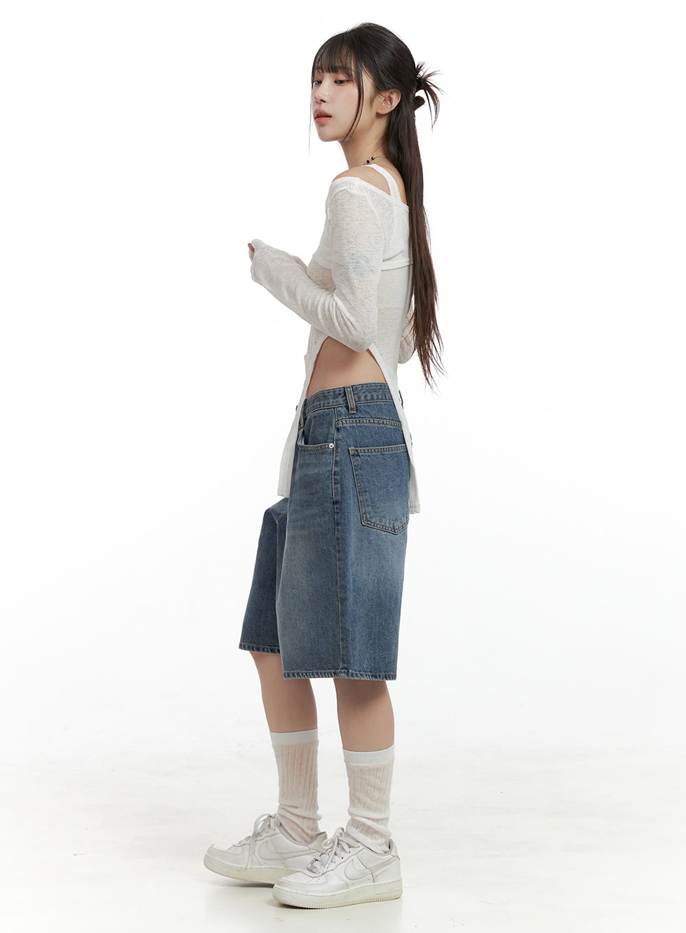 Low Rise Bermuda Jorts CL424 in blue, full-body side profile on model looking back, wide-leg washed denim jorts, back pockets visible, paired with white crop layered top and sneakers.