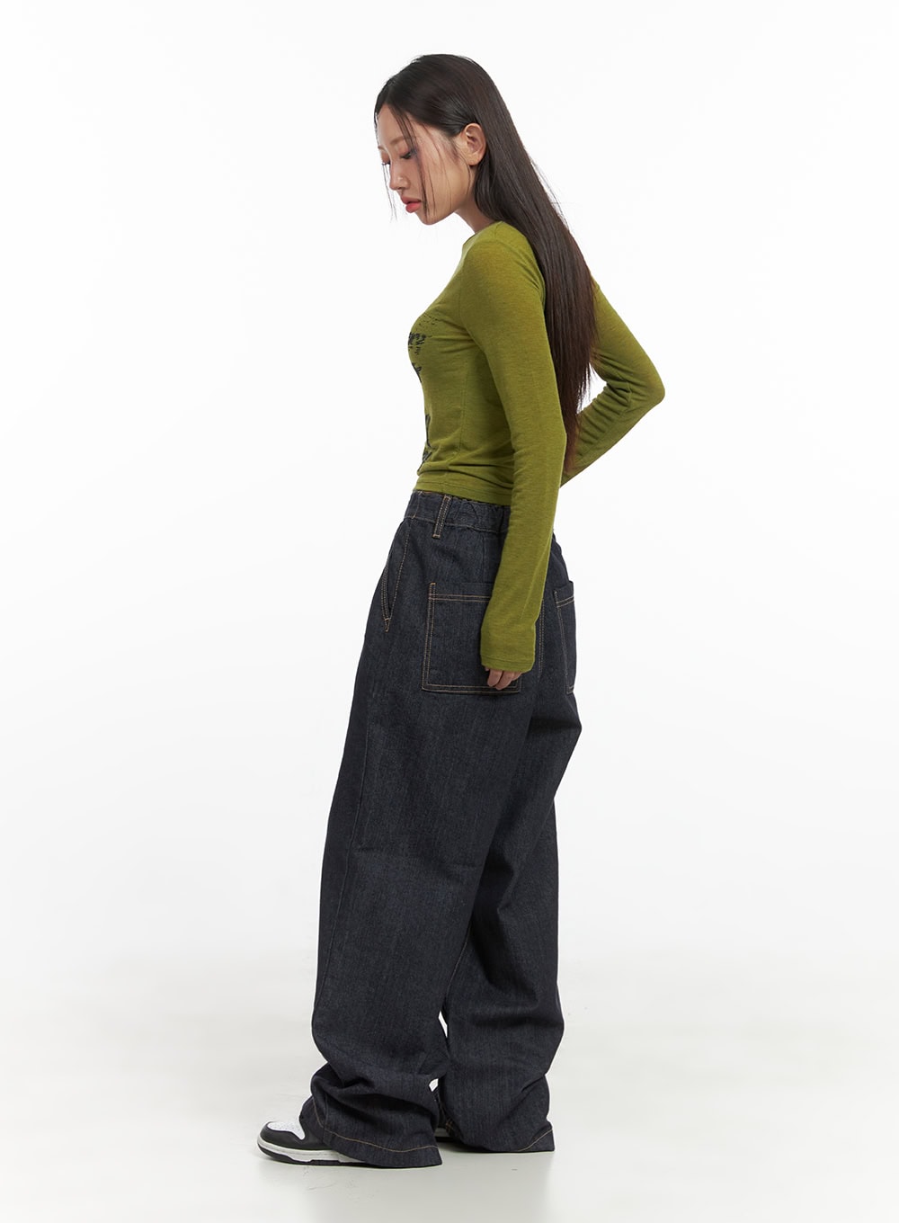 Lola Pintuck Baggy Jeans CO402 in dark blue, side profile on model looking down, showing volume of wide-fit denim leg and contrast topstitching, with olive crop top and sneakers.