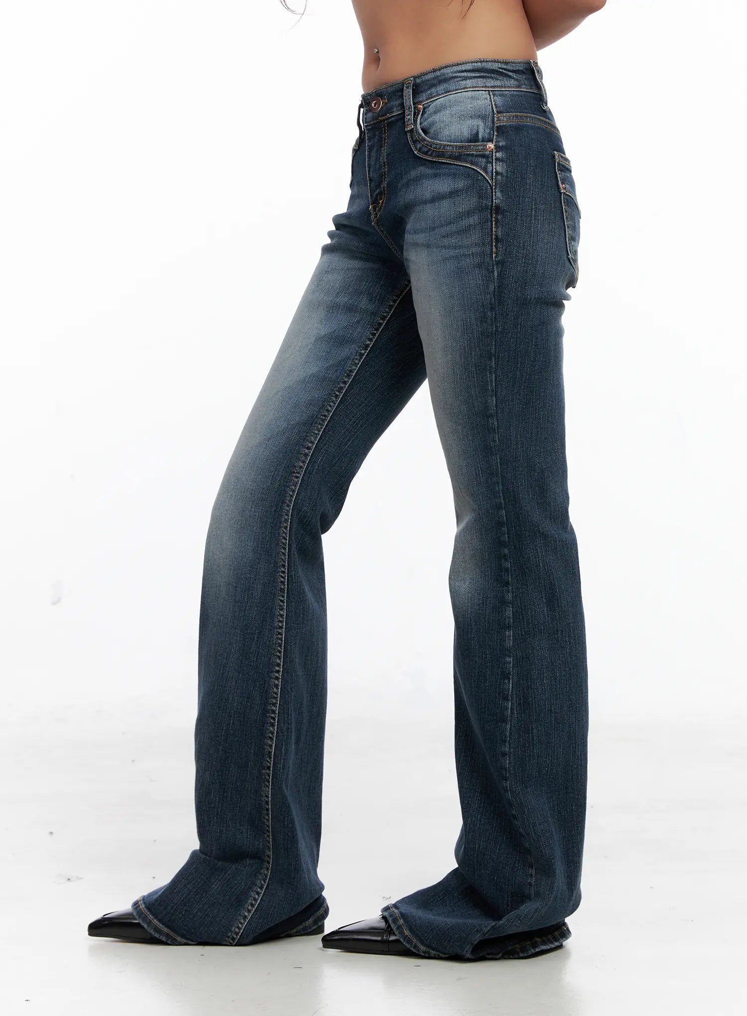 Saanvi Vintage Wash Flare Jeans CS522 in dark blue, side view on model, low-rise waist, bootcut flare hem, curved pocket stitching detail, vintage wash cotton-spandex denim, black pointed flats.