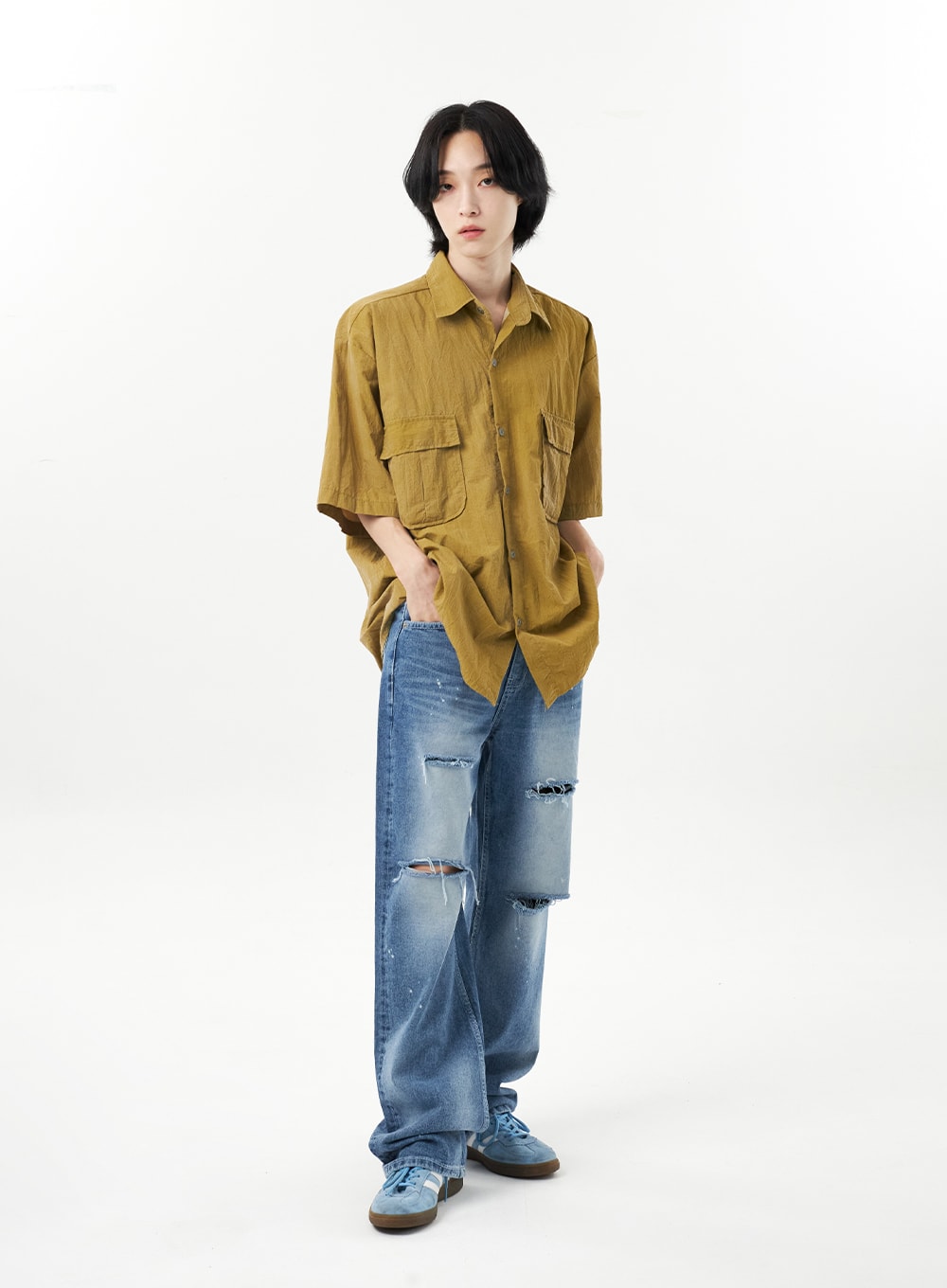 Ripped Jeans Unisex CU315 in mid blue, full-body front on male model, relaxed wide-leg silhouette with distressed knee and thigh rips, paint splatter, worn with mustard utility shirt.
