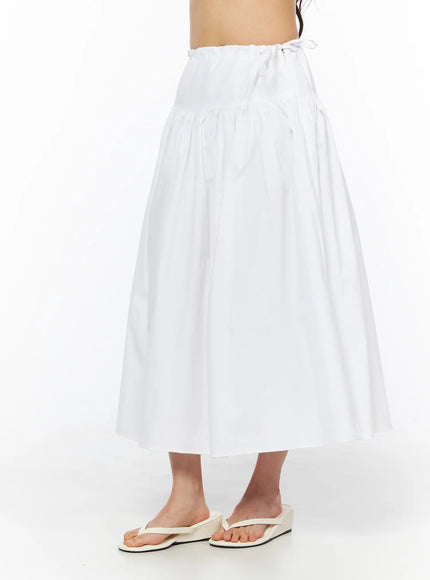 Basic Ribbon Maxi Skirt CU522 in white, side view on model from waist to hem, flared A-line cotton skirt with gathered yoke waist and ribbon tie, worn with white flip-flops.