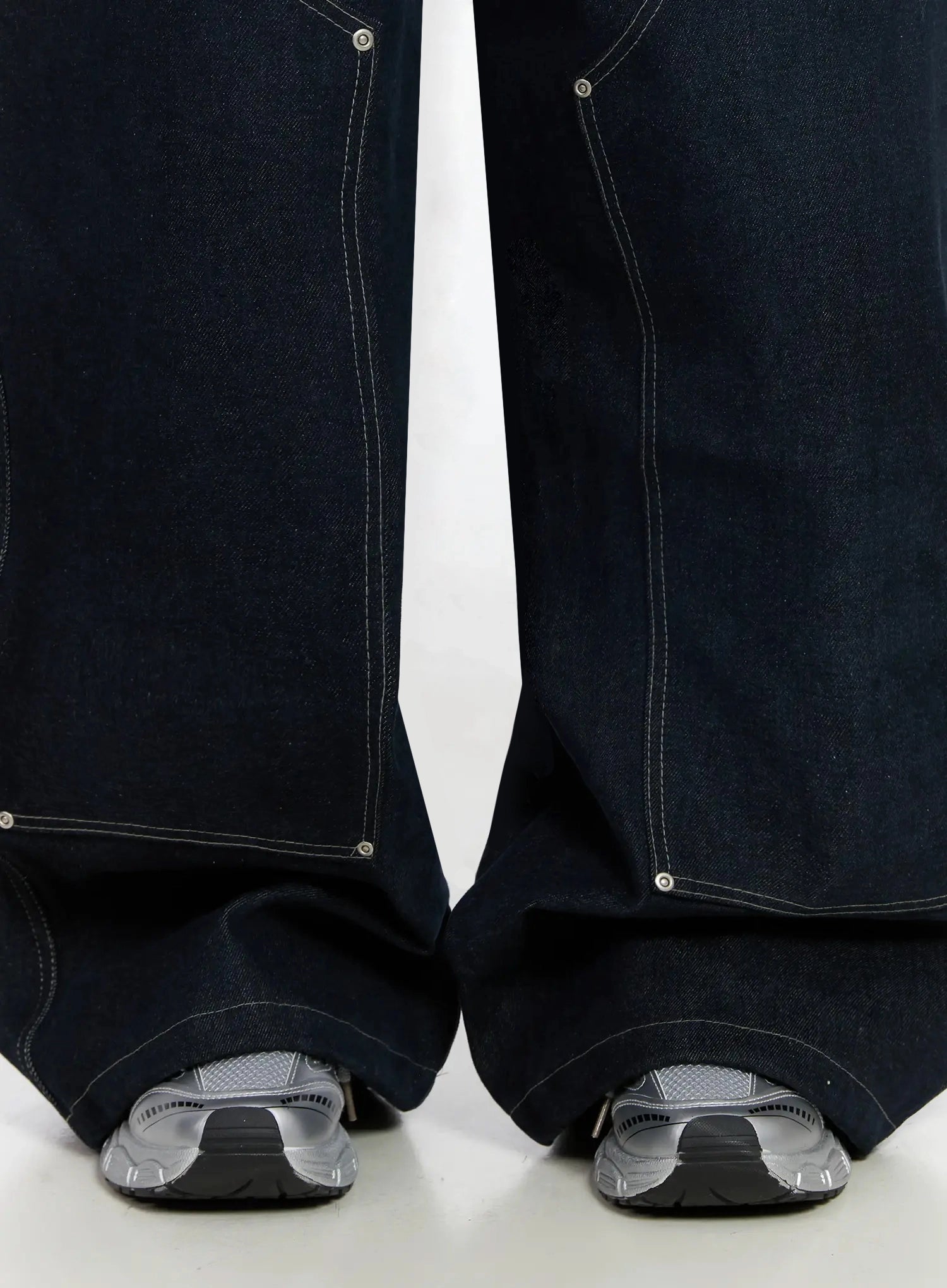 Ashlyn Carpenter Wide-Leg Jeans IJ528 in dark blue, close-up detail of hem and leg opening, contrast white topstitching and silver rivet details, extra-wide pooling hem over silver sneakers.
