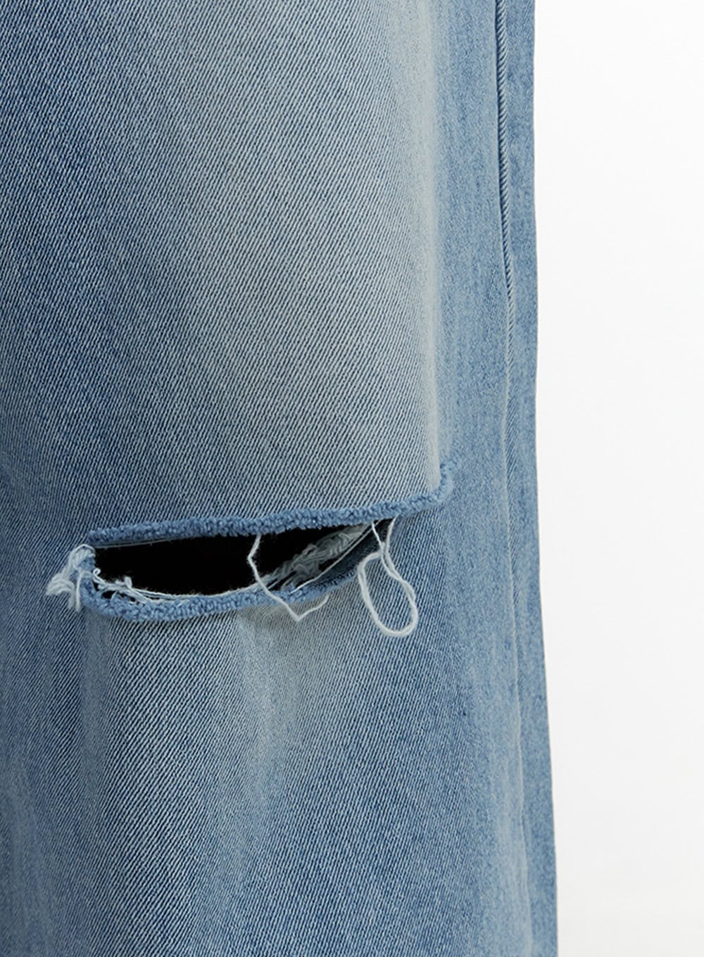 Men's Cut Out Baggy Jeans IU419 in blue, macro close-up of thigh-area cut-out detail showing raw frayed edges and open hole on light wash denim fabric.
