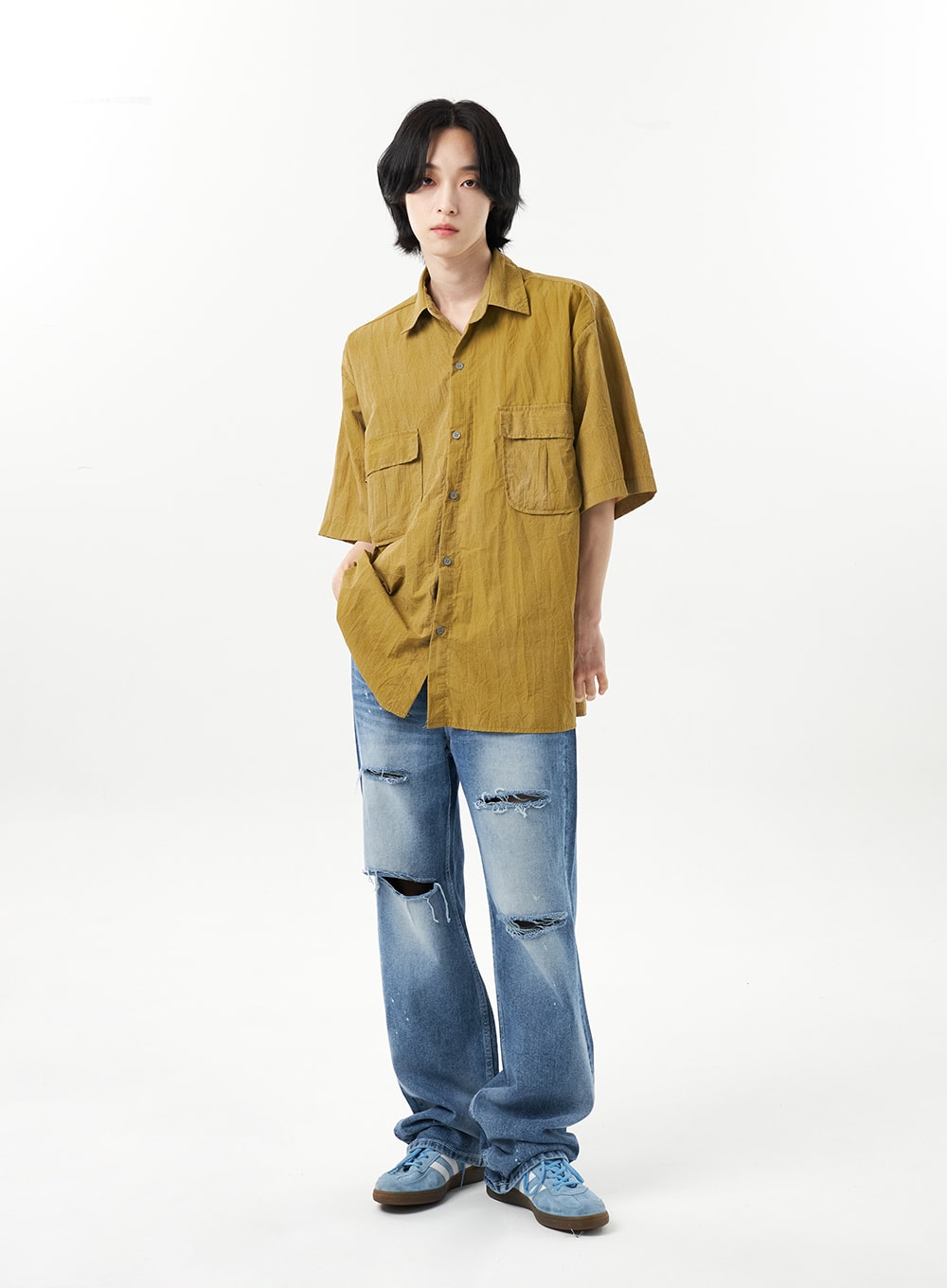 Ripped Jeans Unisex CU315 in mid blue, full-body front on male model, slightly angled stance, wide-fit destroyed denim with visible rips at thigh and knee, styled with olive crinkle shirt.