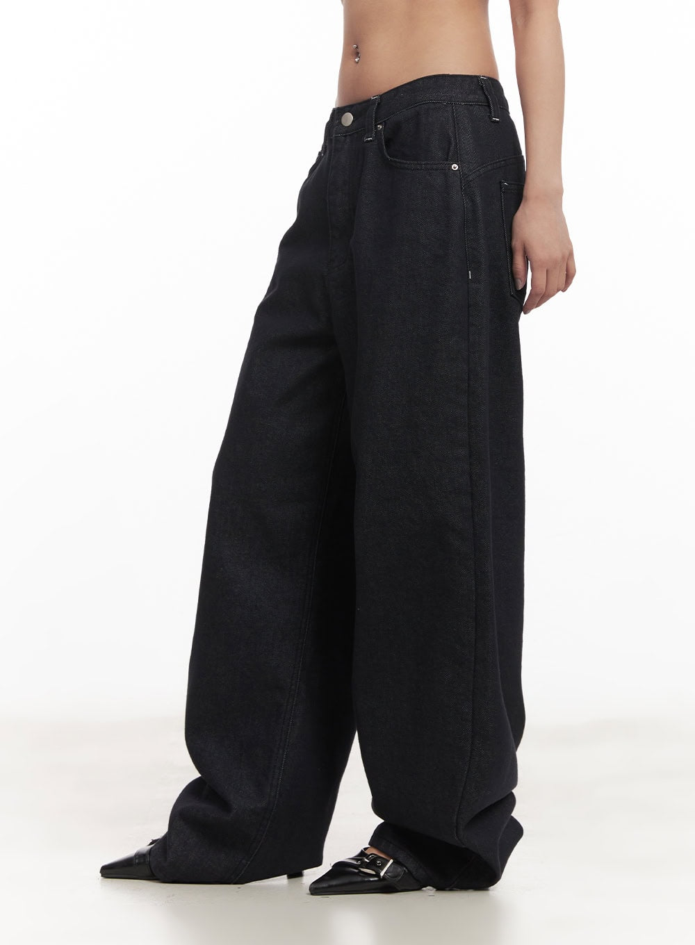 Julianna Baggy Jeans CM505 in black denim, side close-up on model, exaggerated wide-leg and curved hem visible, low-rise waistband with front pockets, worn with black heels.