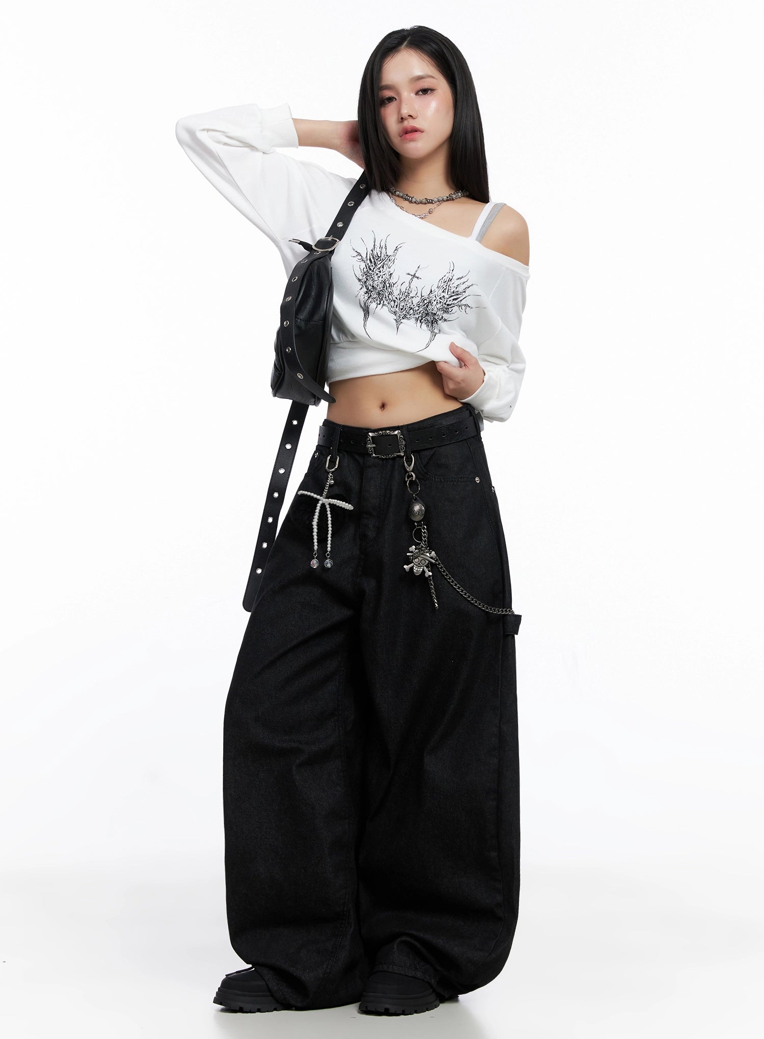 Lea Cross-Patch Baggy Jeans IS525 in black, front full-body editorial shot on model, loose baggy cotton denim styled with chain belt and charm accessories, paired with white graphic off-shoulder top and crossbody bag.
