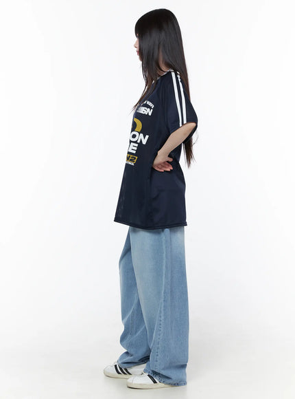 Oversized Sporty Graphic Tee CU519 in dark blue, side view on model, showing drop-shoulder silhouette, white stripe detail on sleeve, mesh fabric, paired with wide-leg jeans and sneakers.