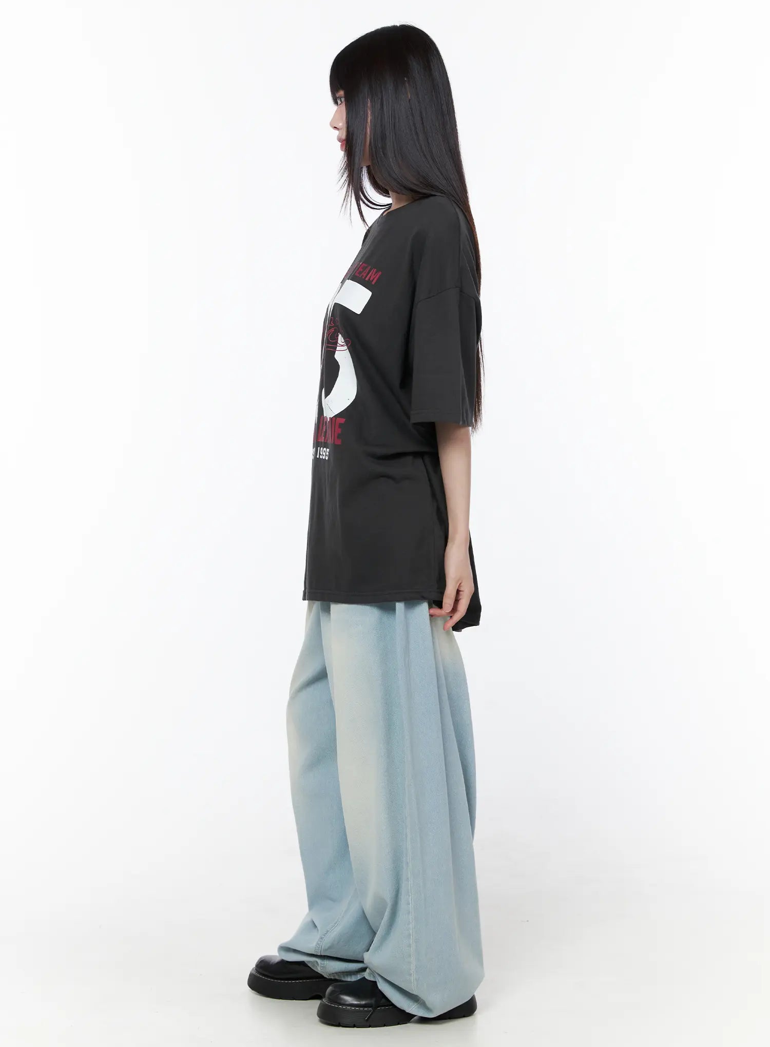 Oversized Graphic Tee CU519 in dark gray, side view on model, showing elongated hem length, dropped shoulders, and curved side seam, paired with light blue wide-leg jeans and black chunky shoes.