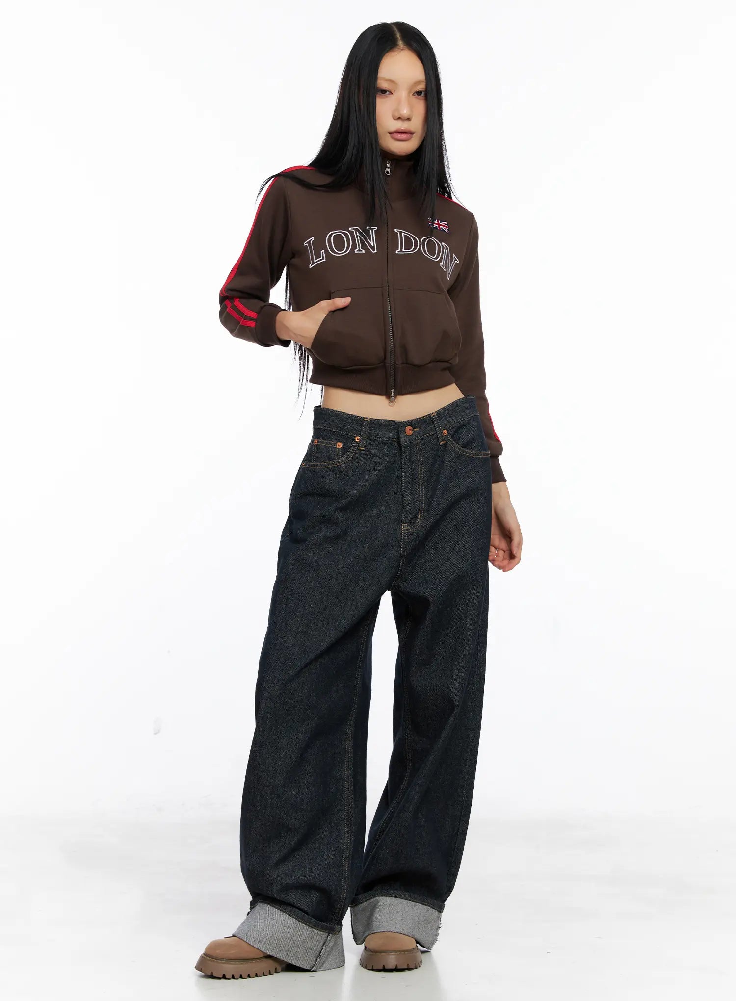 Oakley Cuffed Wide-Leg Jeans CS511 in dark blue, front full-length editorial shot, model standing in brown London graphic crop jacket, loose cotton wide-leg with grey cuffed hem and chunky boots.