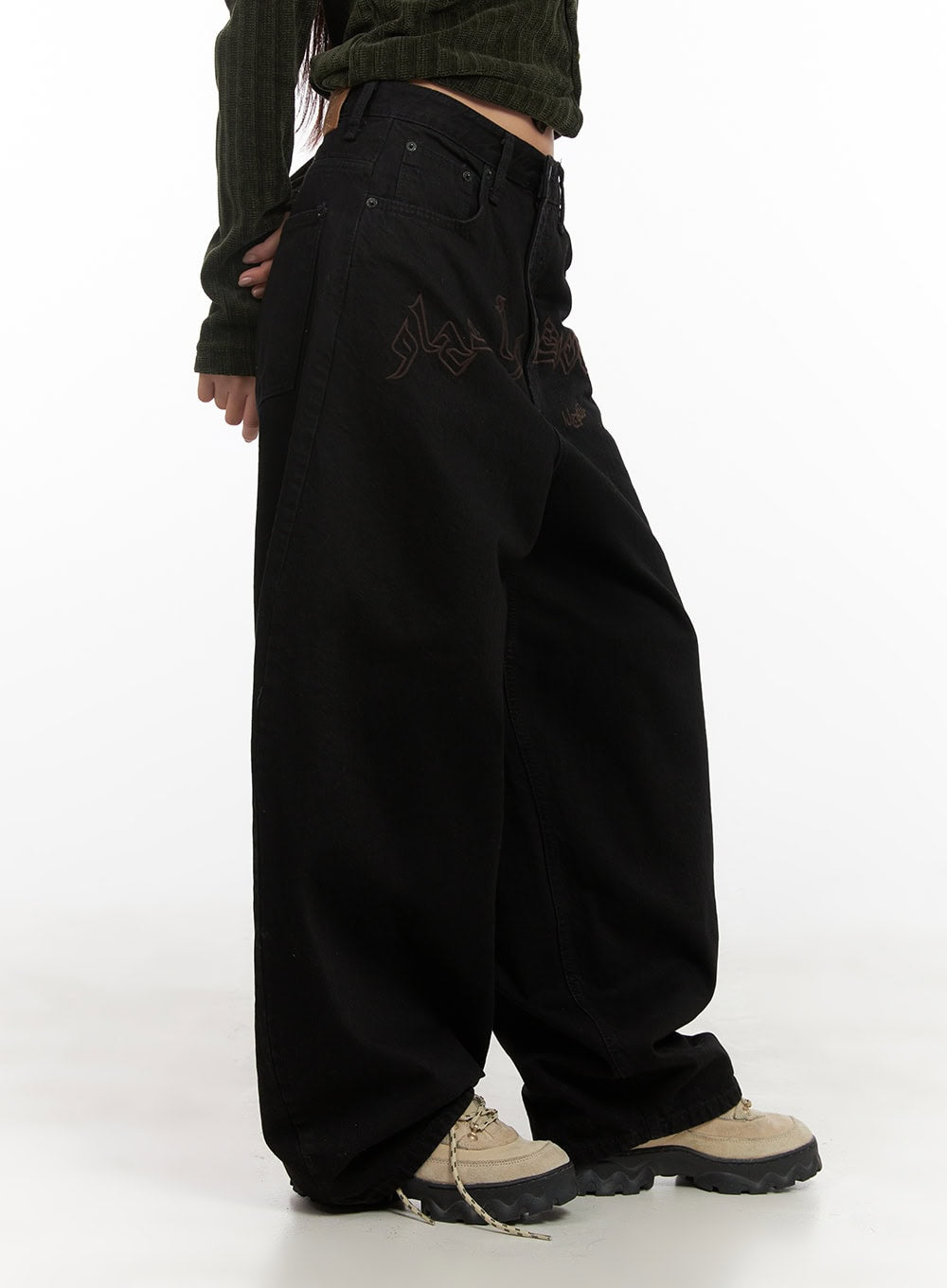 Yasmin Denim Wide Fit Jeans CD413 in black, side close-up on model, dark tone-on-tone calligraphic lettering detail at upper thigh, oversized wide-leg denim silhouette, cropped green knit top above waist.