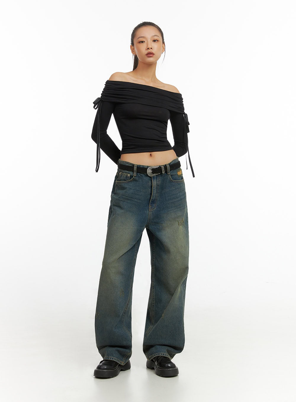 Anya Wide Fit Denim Jeans IO409 in blue, full-length front view on model, baggy washed denim with yellow-tinted fade and distressed texture, paired with black off-shoulder ruched crop top and black western belt.