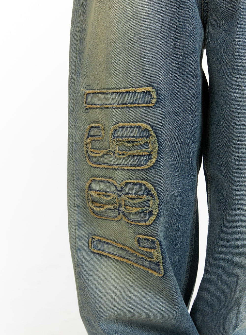 Distressed Baggy Straight Jeans CM413 in blue, macro close-up of '1987' frayed appliqué on left leg, showing raw-edge cutout lettering with yellow-gold stitching on washed denim fabric.