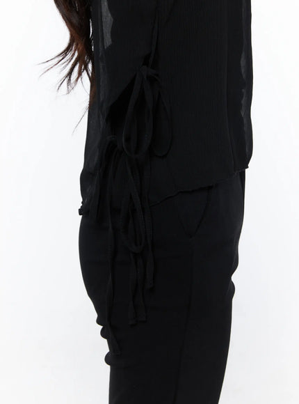 Sheer Ribbon Cami Top CU522 in black, side detail close-up on model, ribbon tie closures at hip, layered sheer hem with crinkle texture, styled with black trousers.
