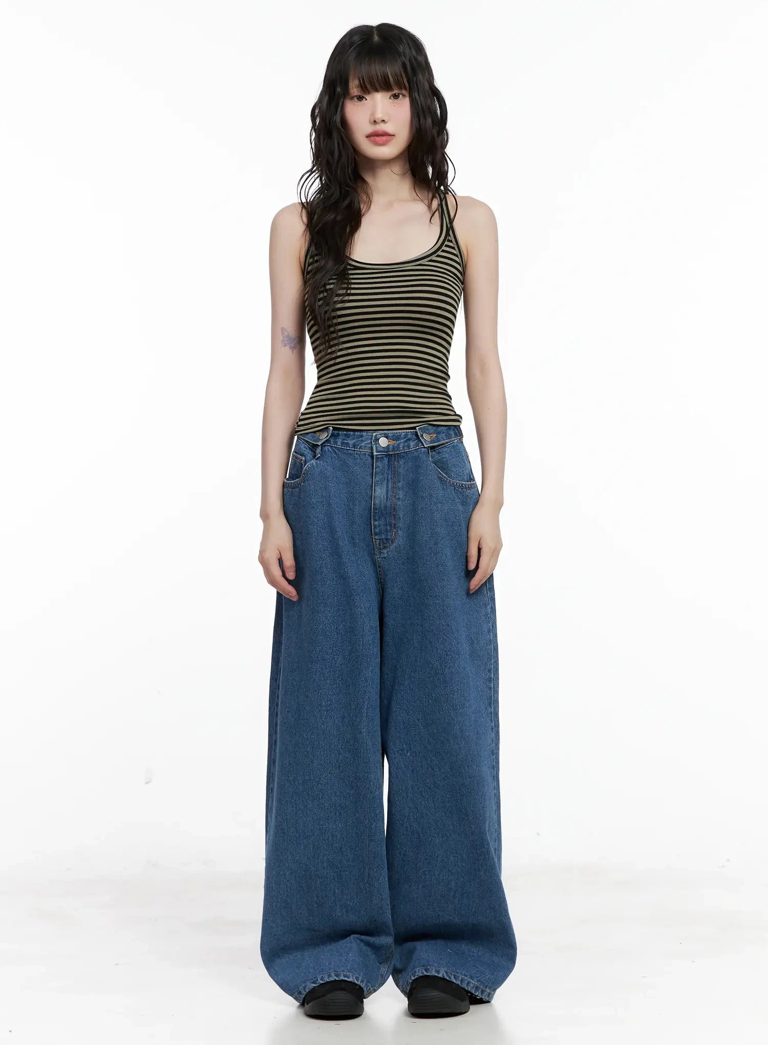 Pharita Loose-Fit Jeans IL528 in mid blue, front full-length view on model, exaggerated wide-leg cotton denim, low-rise crossover waist, paired with olive striped tank top.