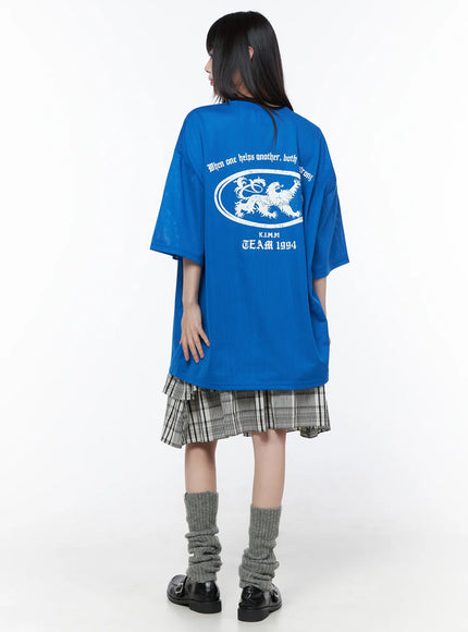 Oversized Mesh Graphic Tee CU519 in Blue M/L, back view on model, white lion crest oval graphic and TEAM 1994 lettering, oversized dropped shoulders, paired with plaid skirt and grey leg warmers.