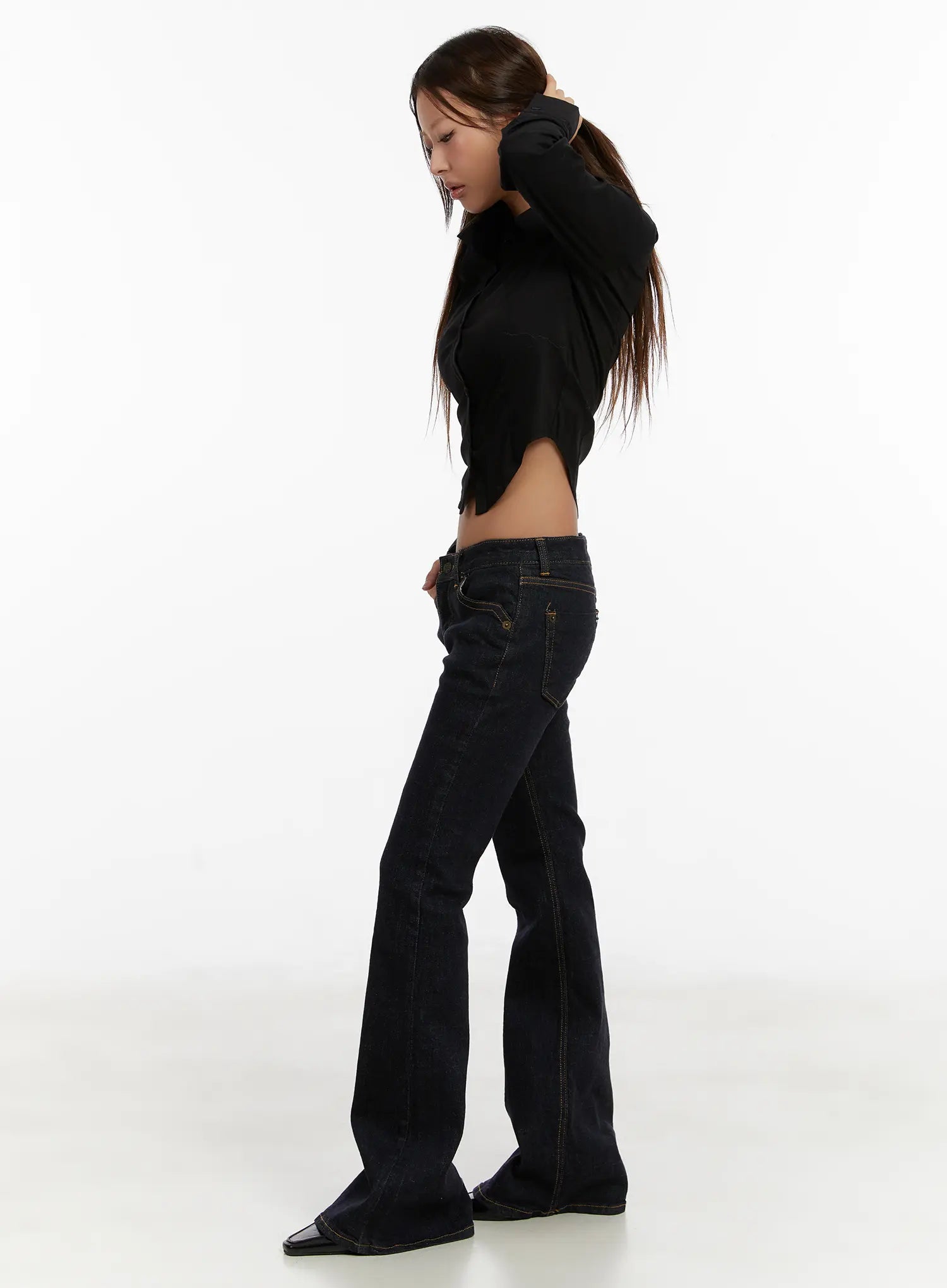Adalee Non-Fade Low-Rise Flare Jeans CD515 in dark indigo, side editorial shot on model in black cropped polo, hand in hair pose, emphasizing low-rise fit and floor-length flare leg.