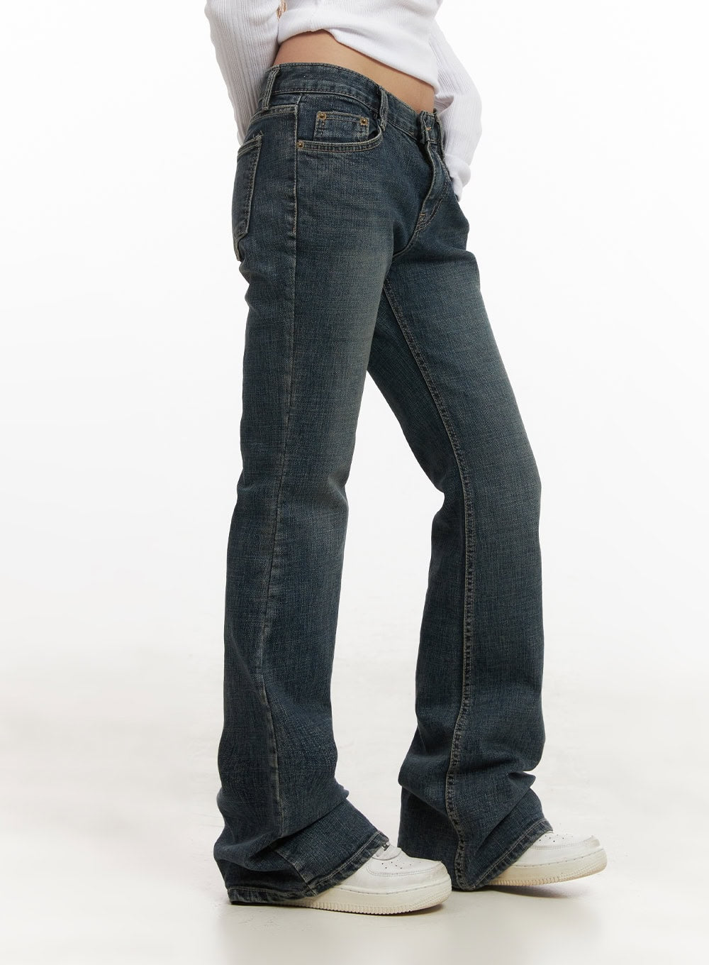 Natalia Low-Rise Flared Jeans CJ508 in dark blue, side close-up on model, washed denim showing front coin pocket, slim fit through thigh and bootcut flare, worn with white ribbed crop top and sneakers.
