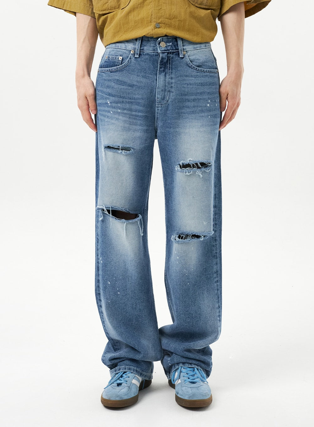 Ripped Jeans Unisex CU315 in mid blue, front waist-to-hem close-up on model, wide-leg denim with large open rips at upper thigh and knee, frayed edges and paint spots visible on cotton fabric.