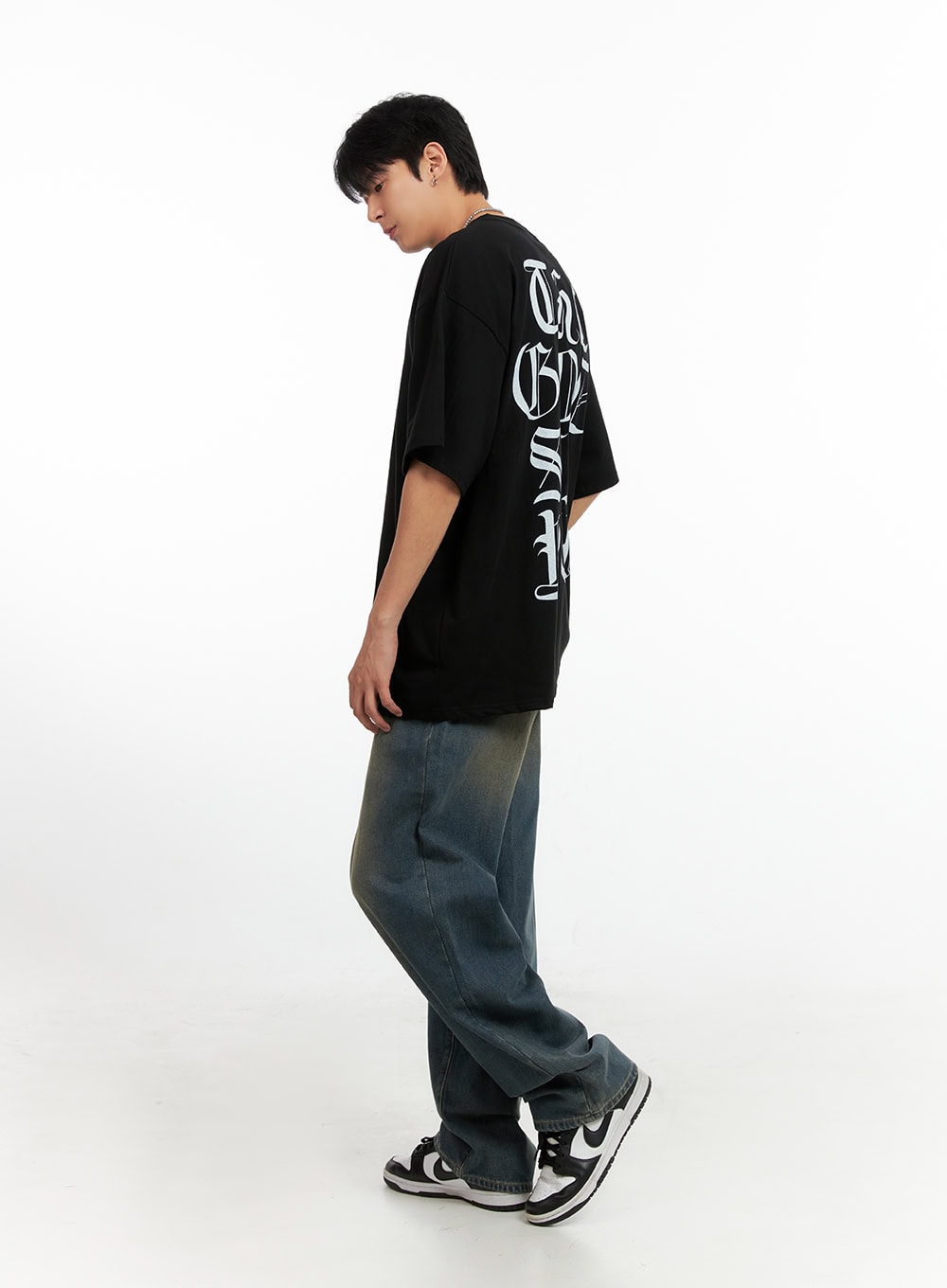 Men's Vintage Washed Loose Fit Jeans (Dark Blue) IG402, side-angle view on model looking down, wide loose leg with warm faded wash visible, styled with black graphic oversized tee and sneakers.