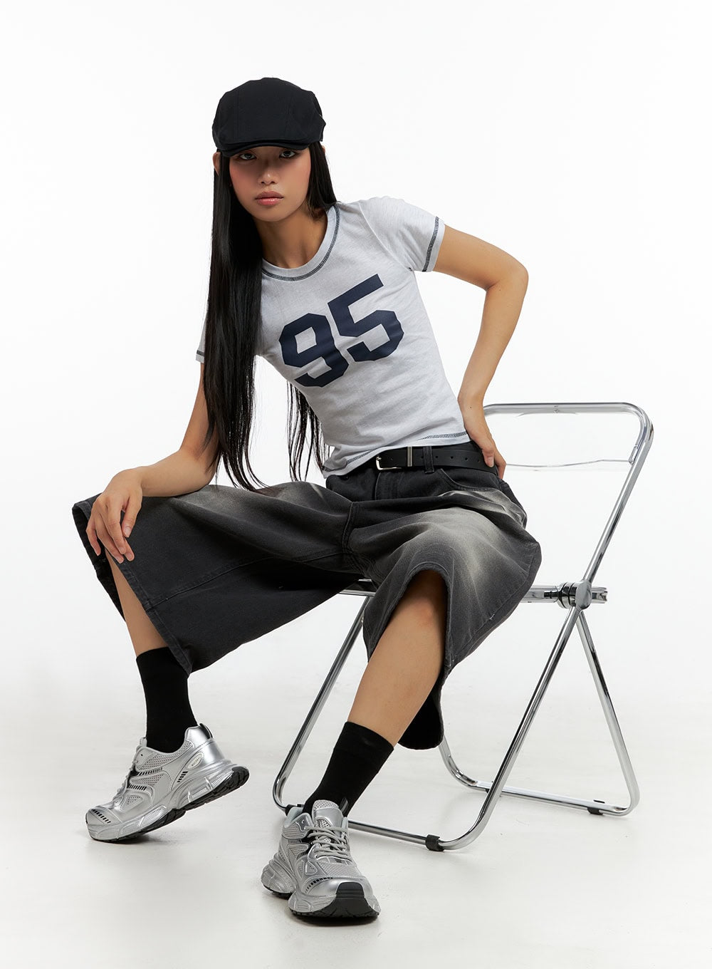 Washed Baggy Jorts IL419 in black, seated editorial shot on model, wide-leg midi denim with washed fade visible, belted waist, paired with crop tee and silver sneakers on chrome chair.
