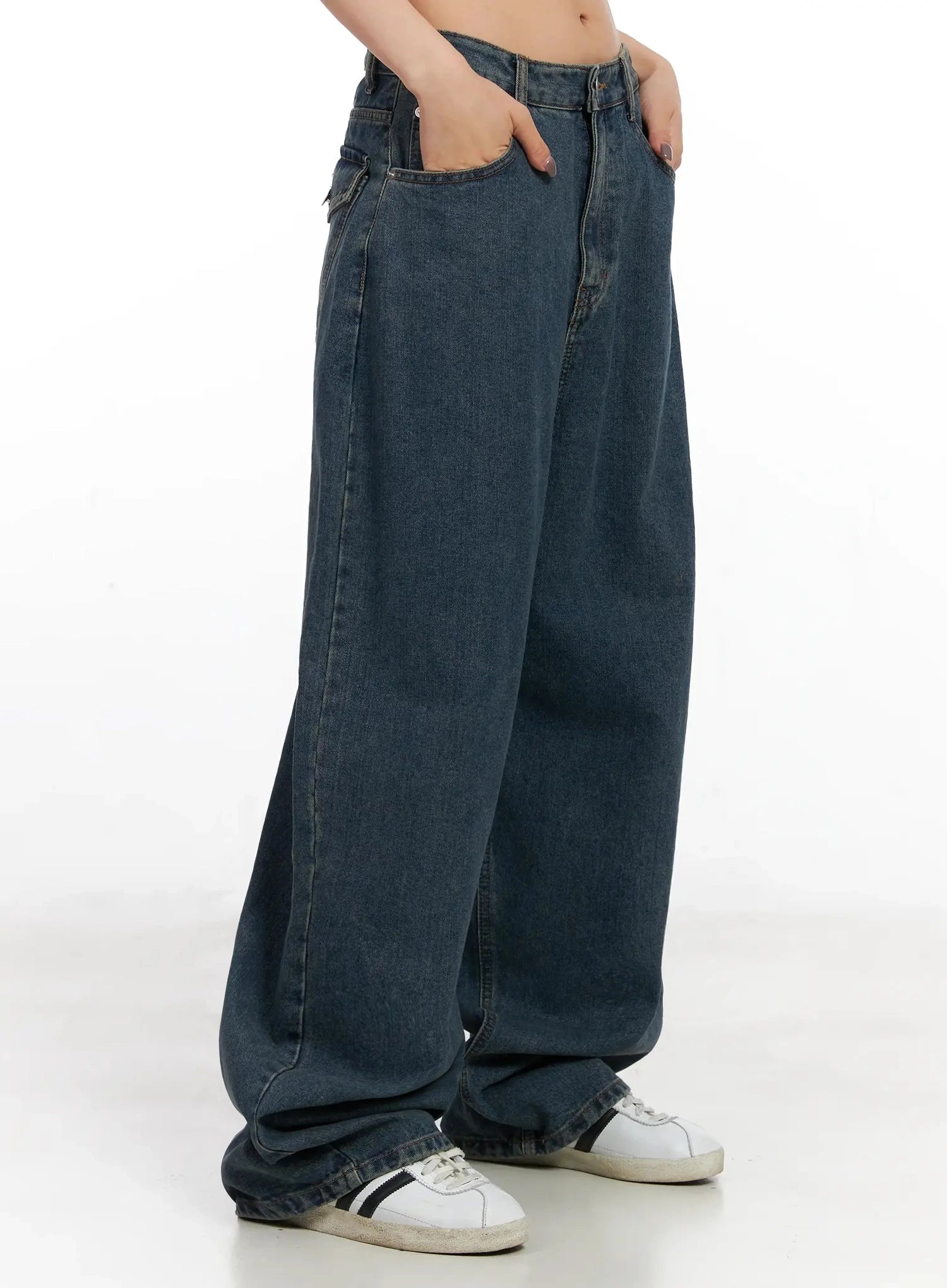 Aleena Mid-Wash Baggy Jeans IN528 in dark blue, side-view shot on model with hand tucked in pocket, showing extreme leg width, long length pooling at hem, worn with white sneakers.