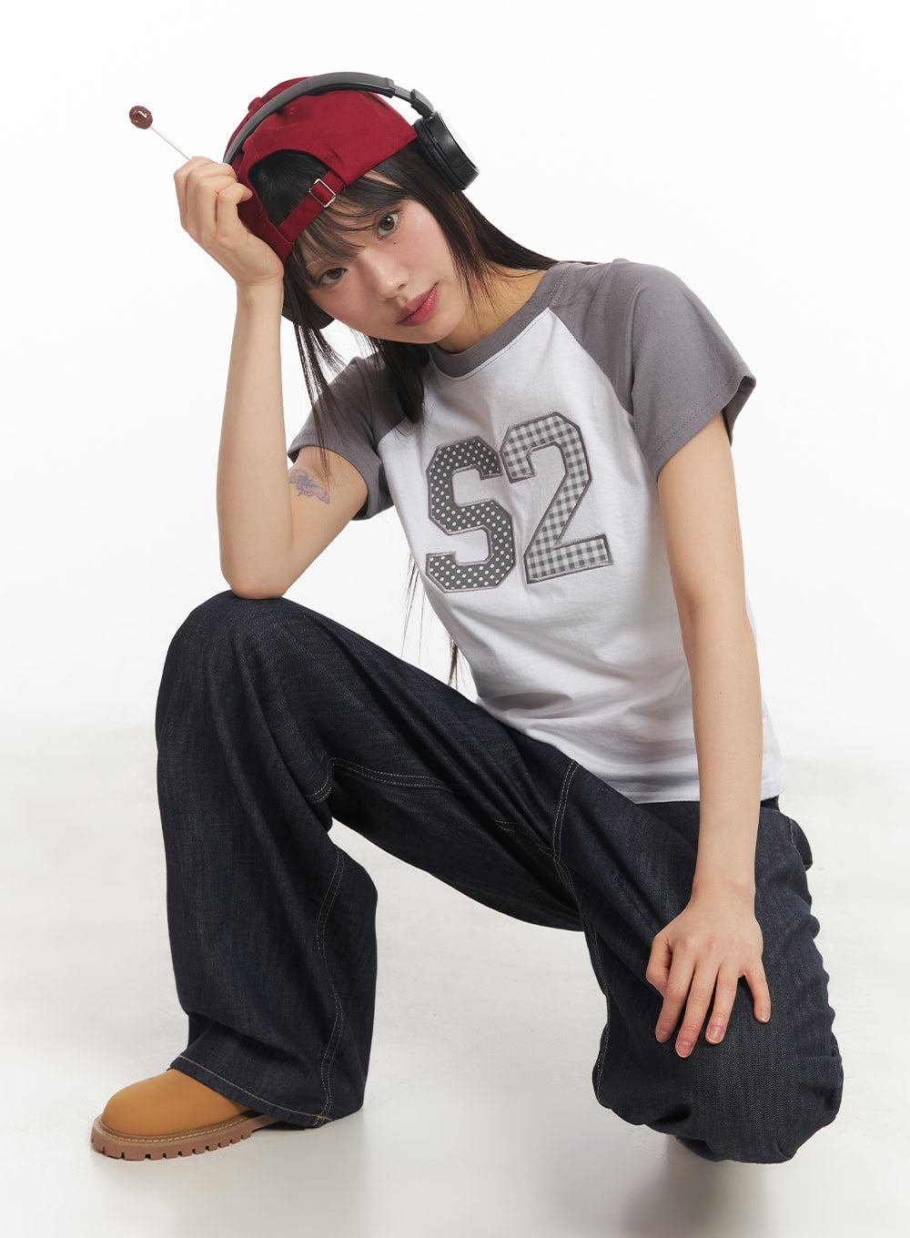 Ivara Wide-Leg Jeans CA522 in dark blue, front three-quarter view on crouching model, wide-leg denim with low rise, paired with graphic raglan tee, red cap, and headphones.