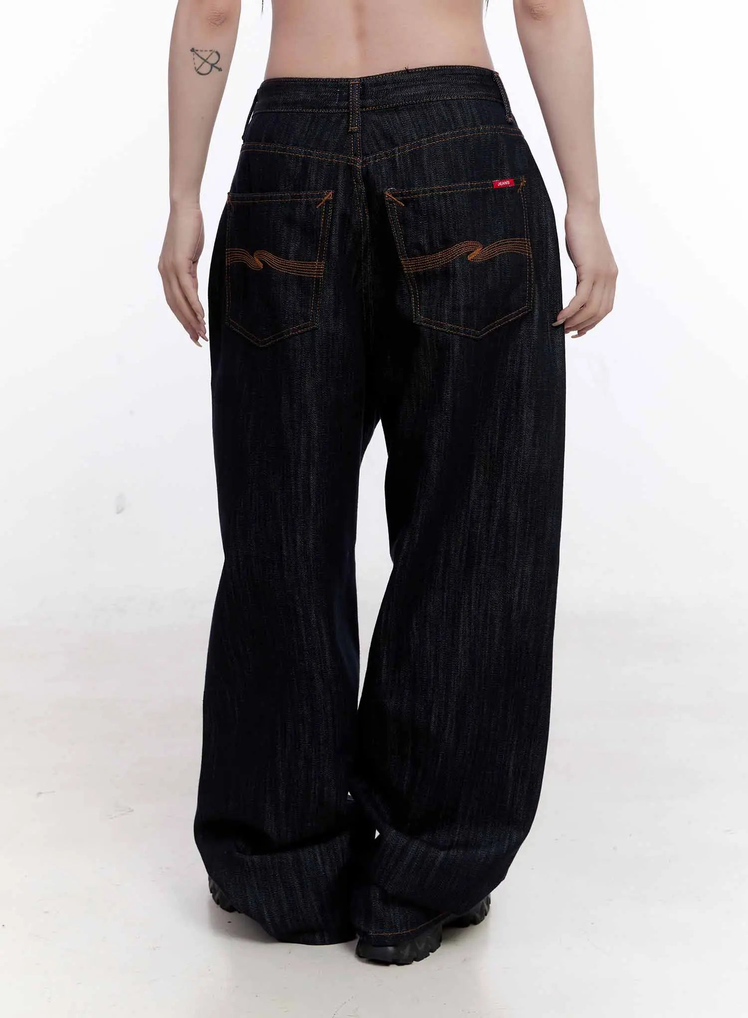 Haewon Dark Washed Baggy Jeans CS530 in dark blue, back full view on model, contrast amber topstitching, back patch pockets with wave embroidery, extra-wide leg hem pooling over sneakers.