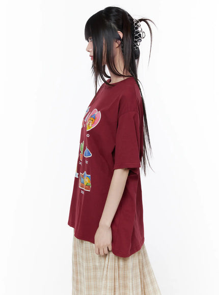 Oversized Graphic Print Tee CU519 in dark red, close side view on model, colorful cartoon sticker prints visible on front, boxy oversized fit with short sleeves, styled over beige plaid skirt.