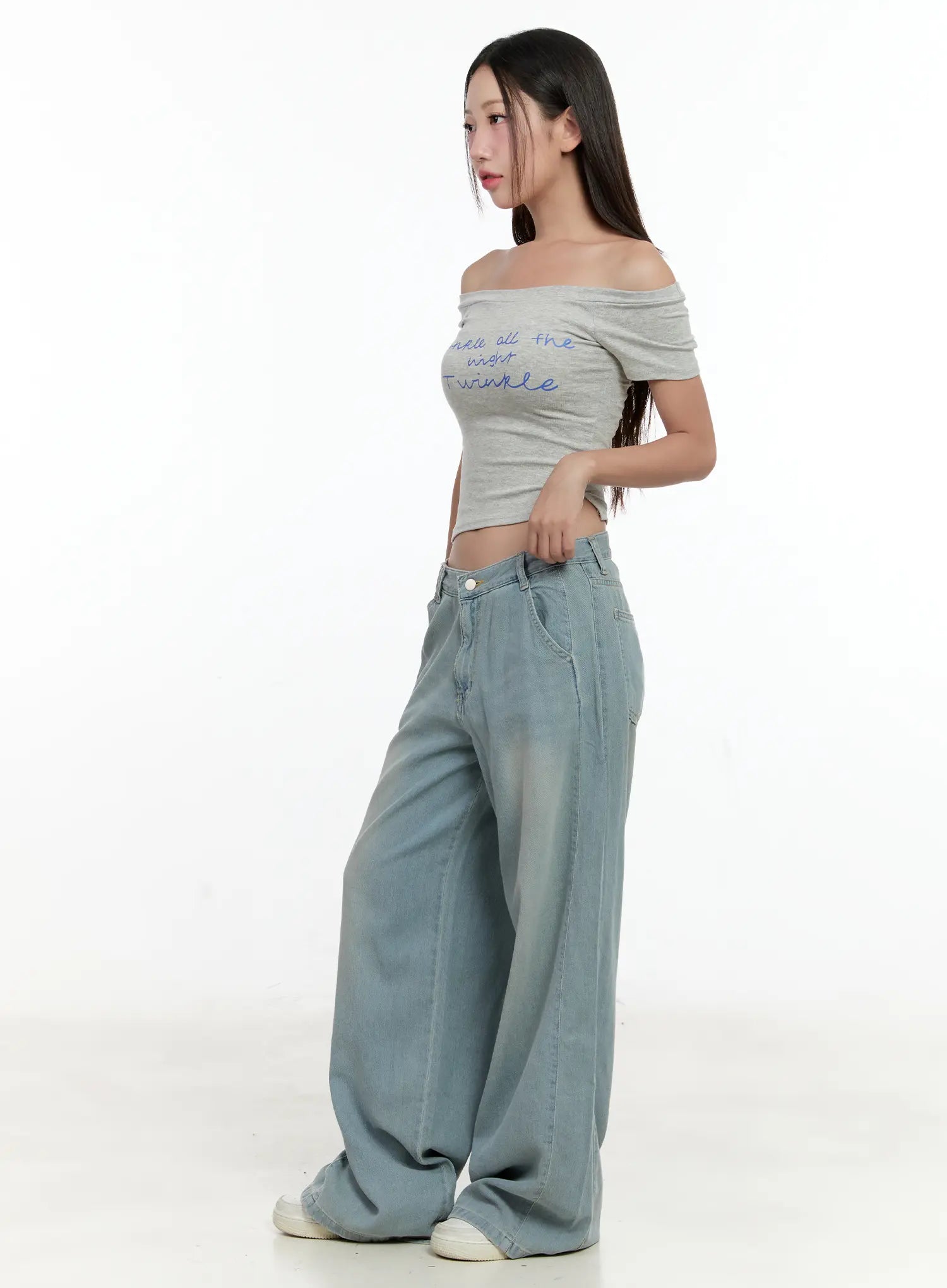 Huening Wide-Leg Jeans CL531 in blue, side view on model in profile pose, washed wide-leg denim with low-rise V waistband, styled with grey off-shoulder crop top and white sneakers.