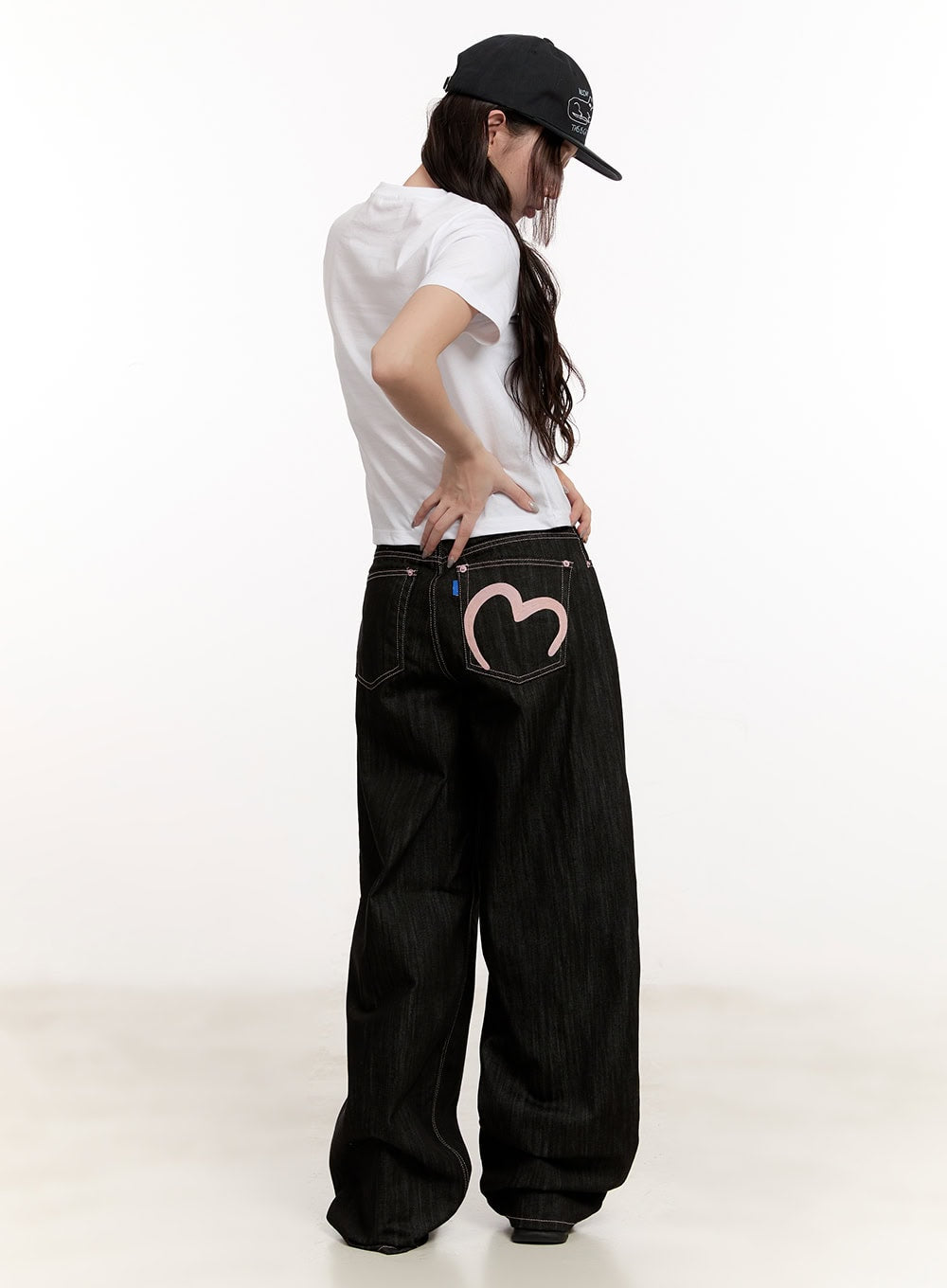 Trinity Stitched Baggy Jeans CM510 in black, back view on model, large pink heart embroidery across back pocket, wide baggy leg, white tee and black cap styling.