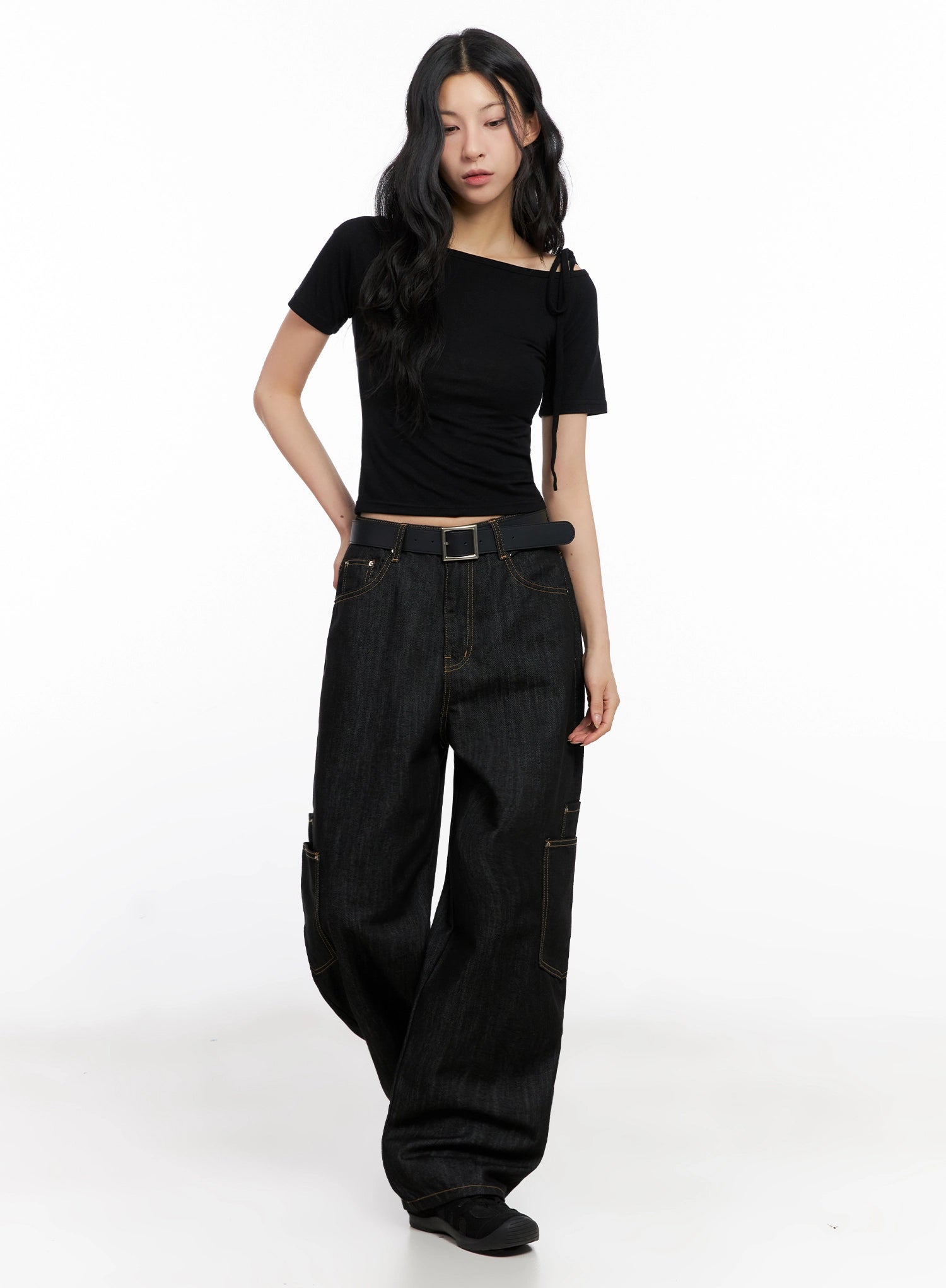 Dahee Wide-Leg Cargo Jeans IG508 in black, full-body three-quarter angle on model looking down, belted at waist with black strap, wide-leg cargo silhouette with contrast gold stitching, paired with sneakers.