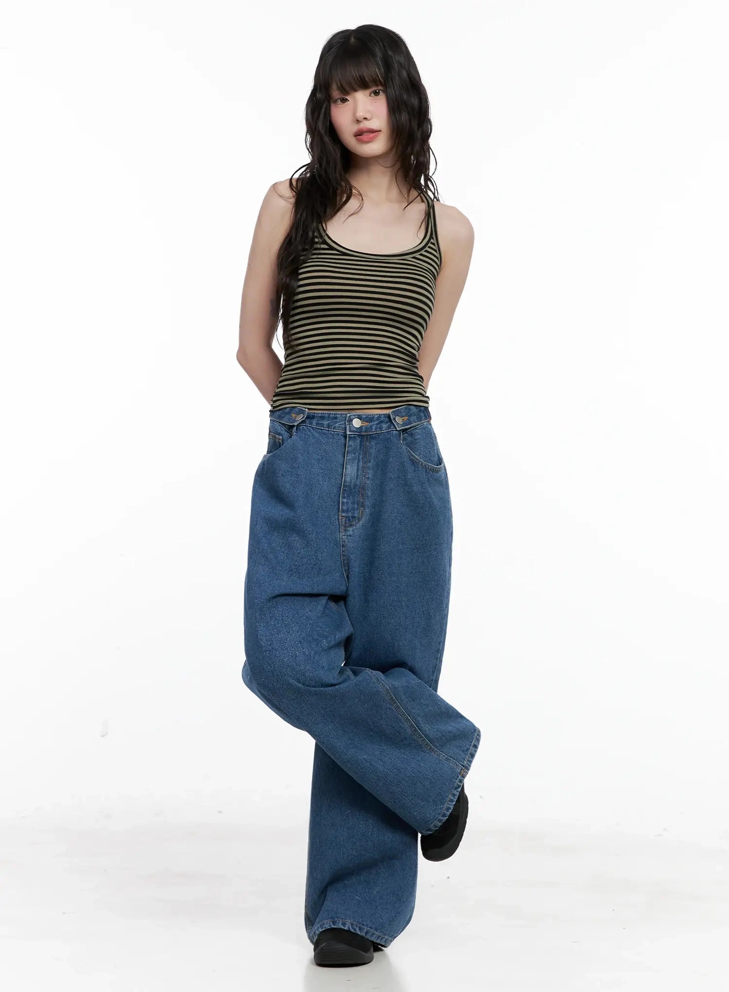 Pharita Loose-Fit Jeans IL528 in mid blue, three-quarter front view on model with one leg raised, wide-leg cotton denim showing voluminous drape, styled with olive striped tank top.