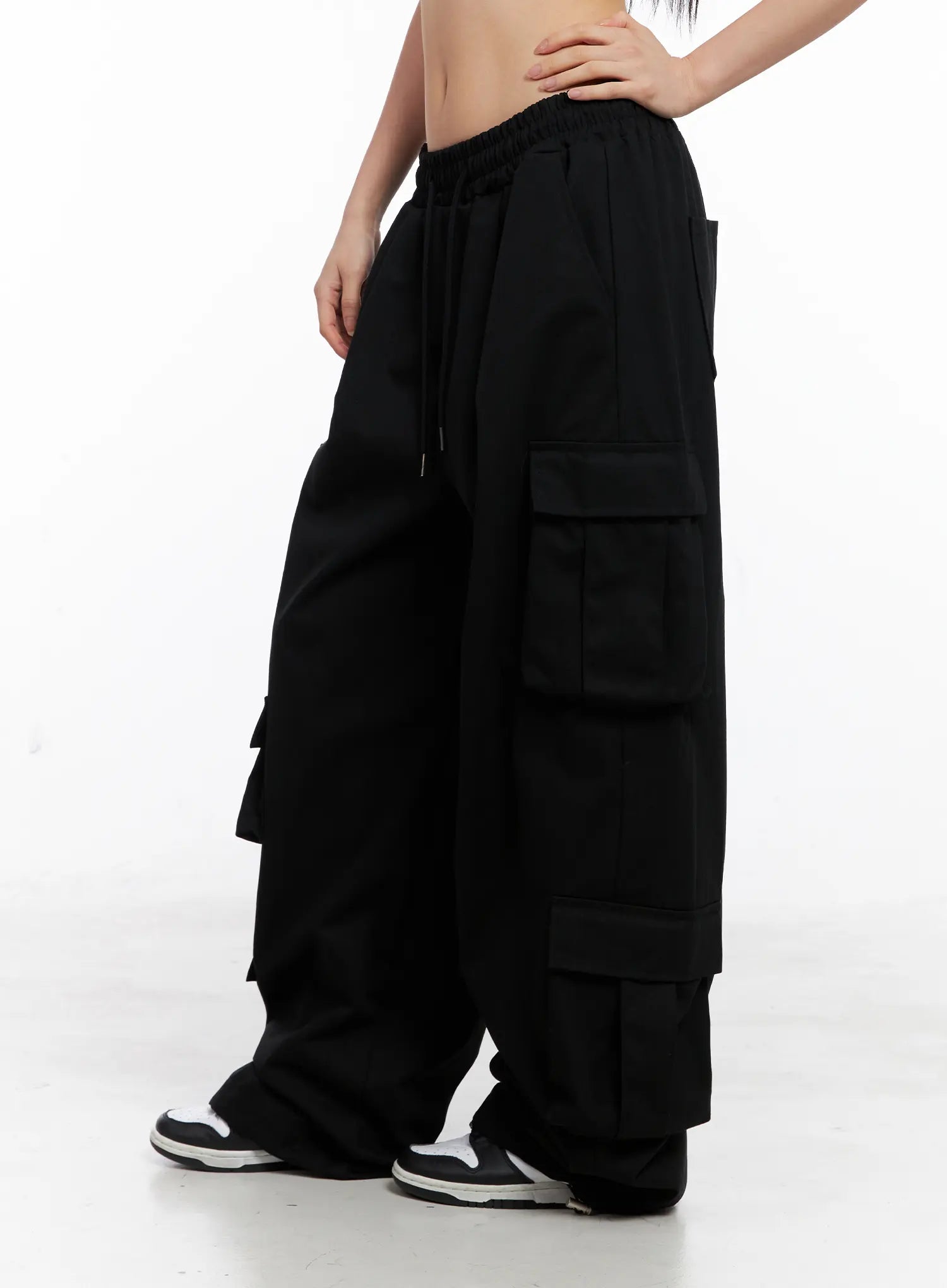 Wide Leg Cargo Pants CC1109 in black, side mid-shot on model, hand on waist, elastic drawstring waist, layered cargo pockets on thigh and lower leg, pooling at sneakers.