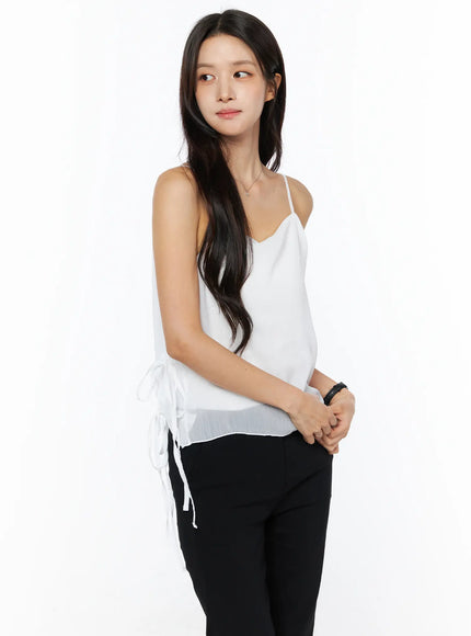 Sheer Ribbon Cami Top CU522 in white, three-quarter side view on model, V-neck thin straps, side ribbon ties at hem, sheer crinkle fabric, paired with black trousers.