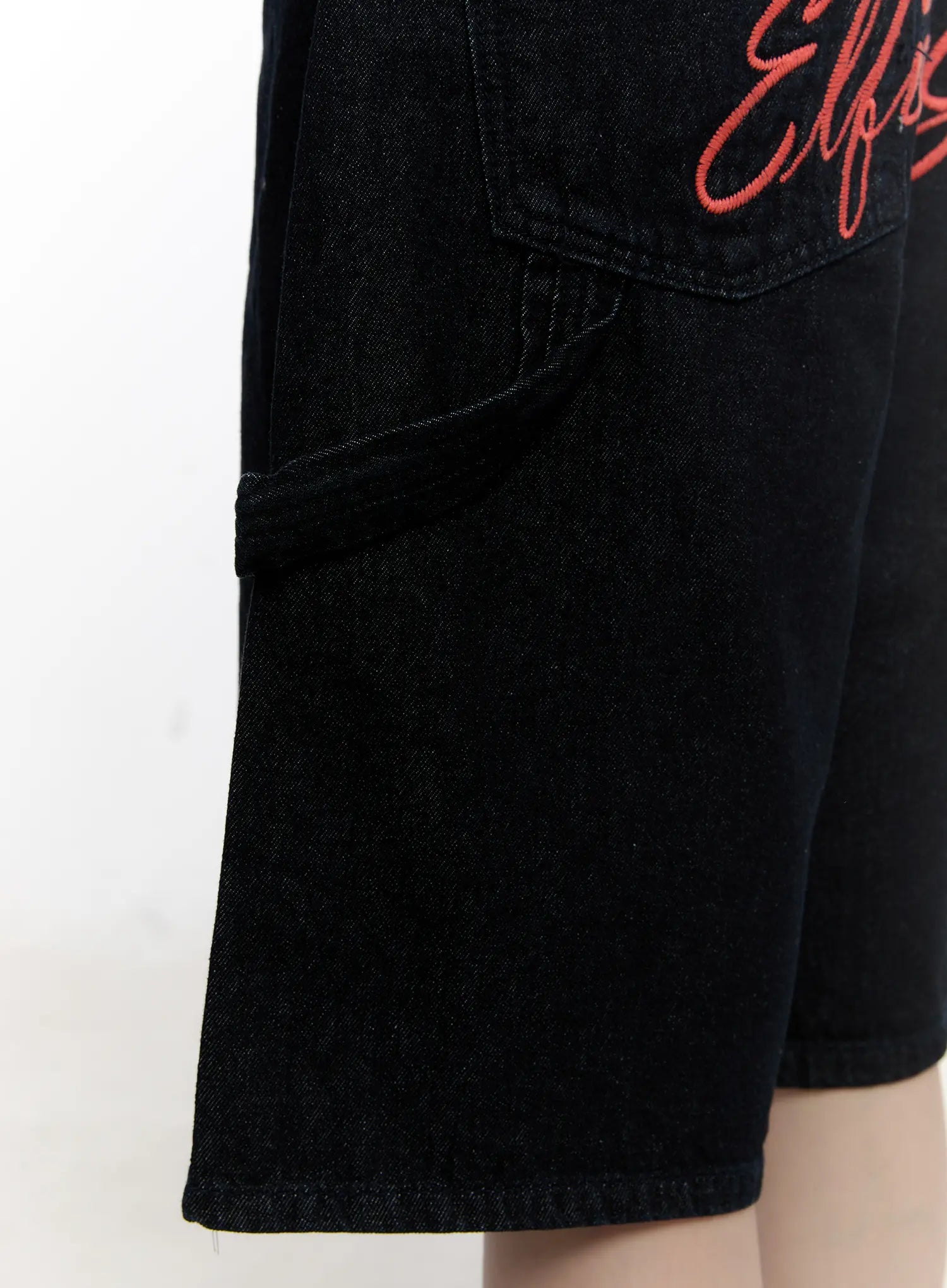 Red Text Graphic Embroidered Jorts CL504 in black, detail close-up of side utility pocket with belt-loop strap, red script embroidery above, washed cotton denim texture.