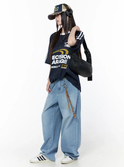 Oversized Sporty Graphic Tee CU519 in dark blue, three-quarter editorial shot on model with cap and headphones, oversized mesh tee with racing graphic, styled with wide-leg jeans and shoulder bag.
