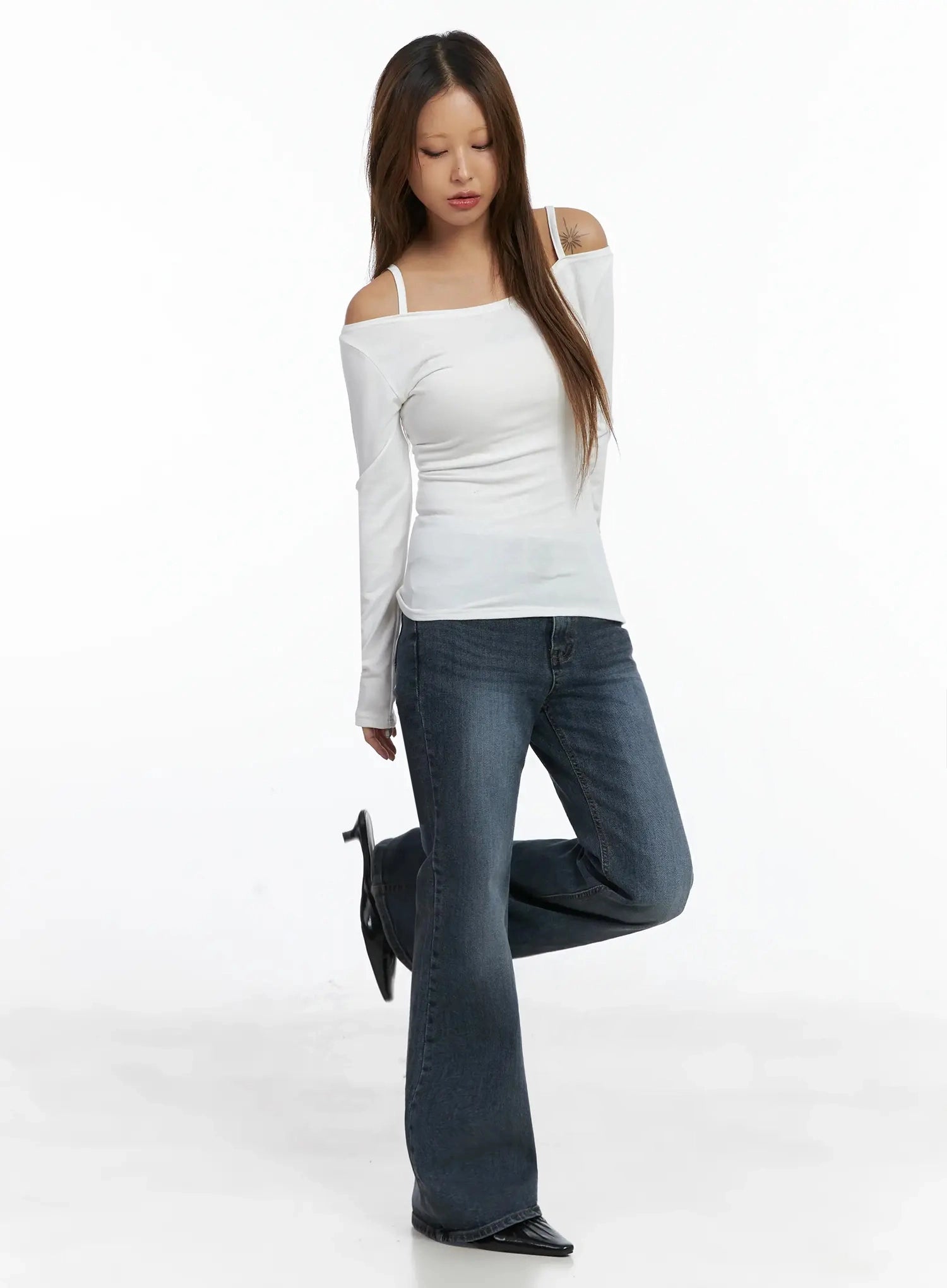Rara Fleece Flared Jeans ID503 in blue, full-body angled pose on model with one leg raised, washed flared hem pooling at foot, paired with white off-shoulder top and black heeled mules.