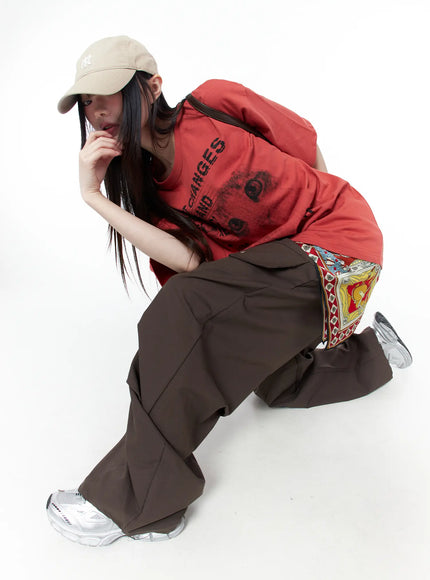 Oversized Cat Print Graphic Tee CU519 in dark red, dynamic crouching editorial pose on model with NY cap, cat graphic visible on chest, layered with patterned scarf and brown wide-leg cargo pants.