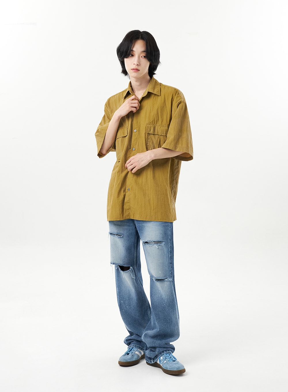 Ripped Jeans Unisex CU315 in mid blue, full-body front on male model with hand raised to collar, wide-leg destroyed denim with thigh and knee rips, paint splatter, styled with mustard shirt.