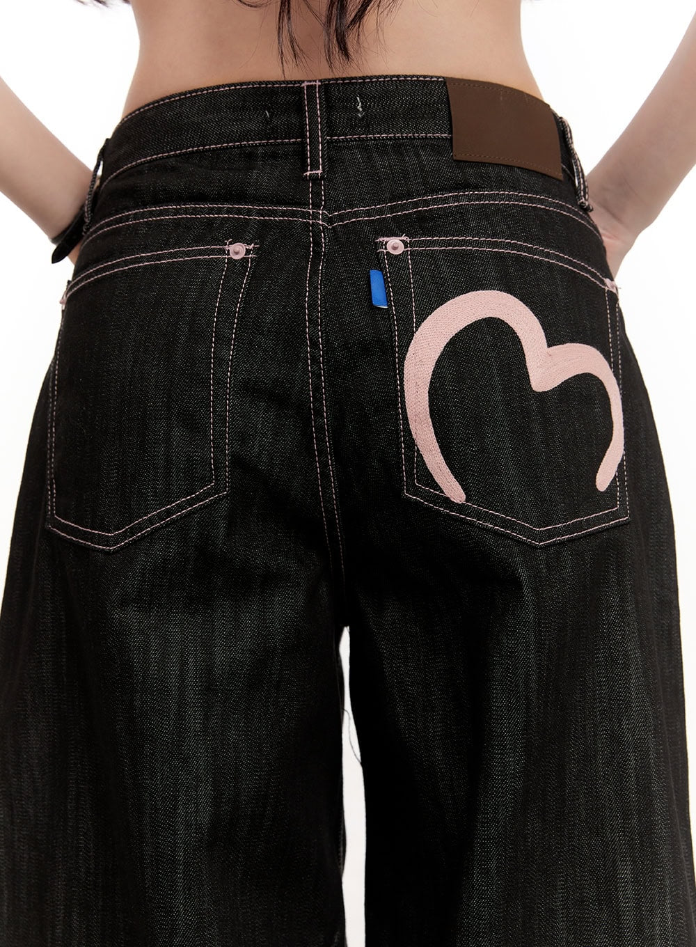 Trinity Stitched Baggy Jeans CM510 in black, close-up back detail shot, oversized pink heart embroidery on right back pocket, contrast stitching, blue tab, brown leather label.