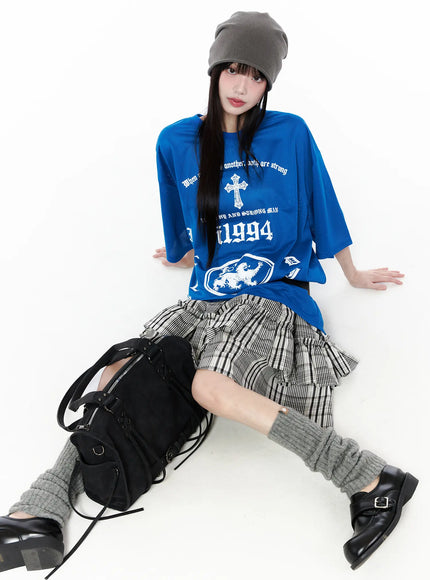 Oversized Mesh Graphic Tee CU519 in Blue M/L, seated editorial shot on model with grey beanie, front gothic cross and i1994 lion prints showing, styled with plaid ruffle skirt and black bag.