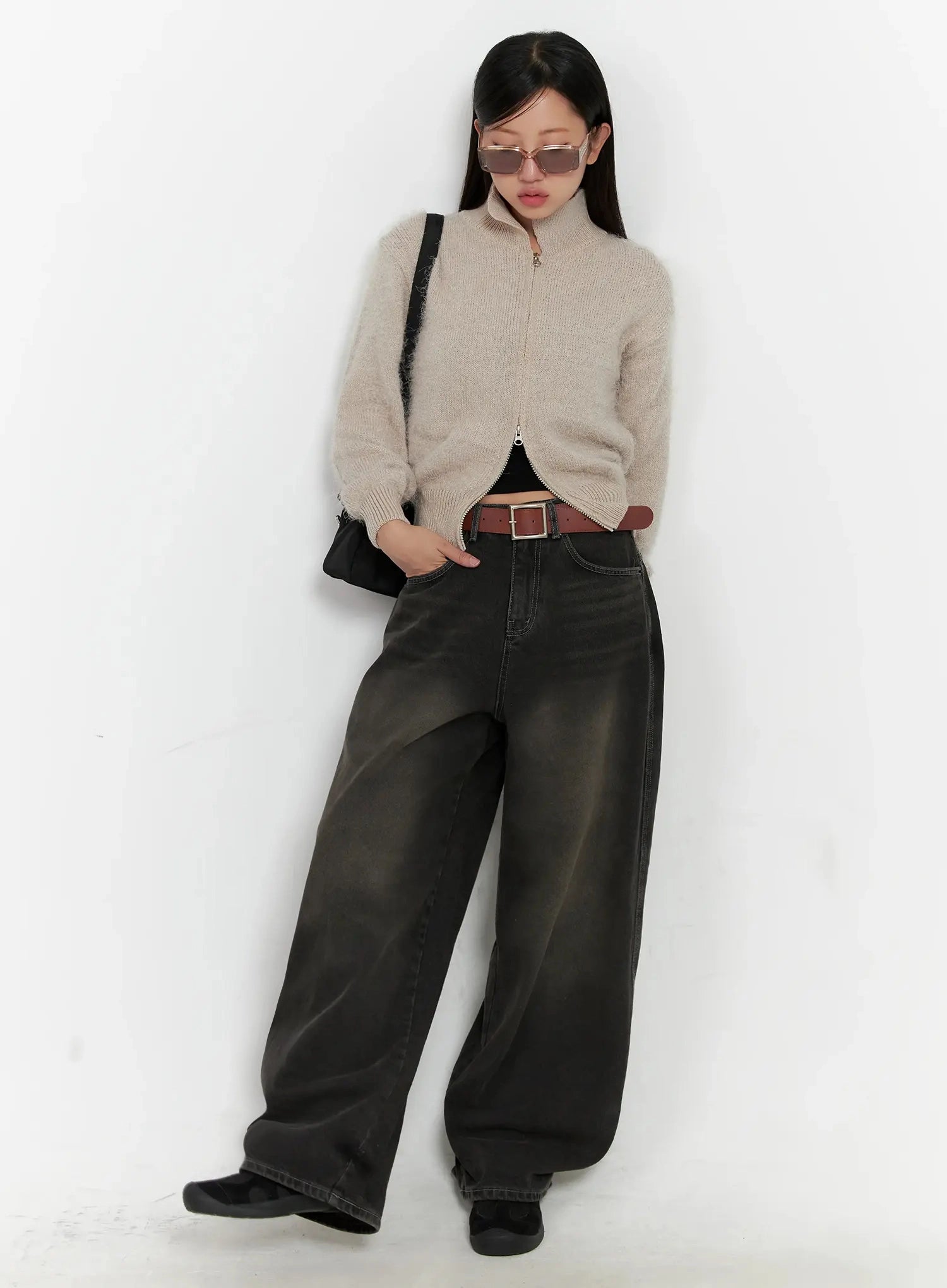 Hari Dark Washed Baggy Jeans IG527 in dark grey, full-length editorial shot on model leaning against wall, styled with beige zip knit, brown belt, sunglasses and black shoulder bag.