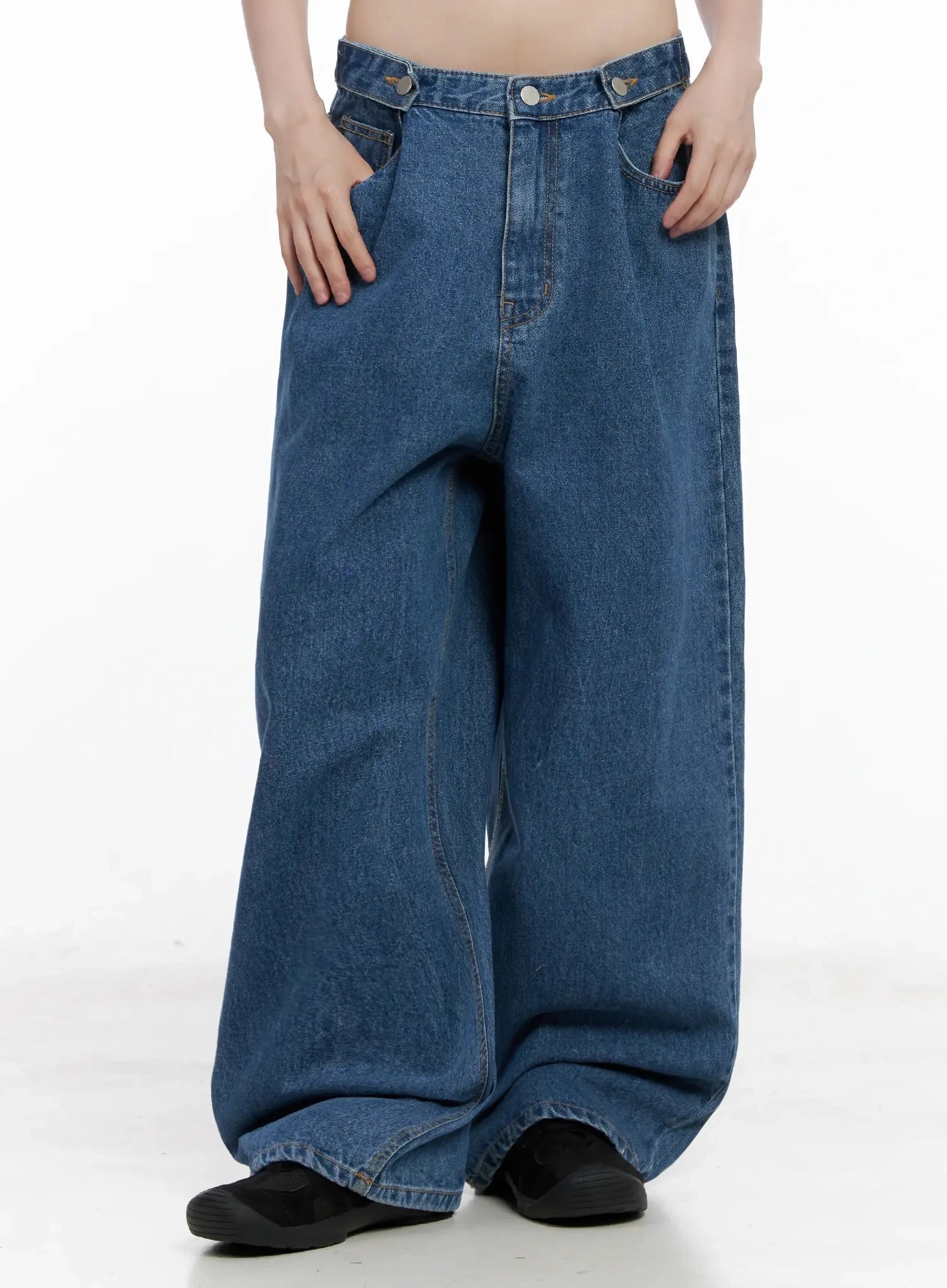 Pharita Loose-Fit Jeans IL528 in mid blue, front bottom-half shot on model, extra-wide leg opening with pooled hem, asymmetric snap waistband detail, hands in pockets.