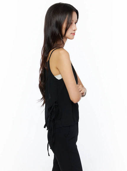 Sheer Ribbon Cami Top CU522 in black, full side profile on model, thin spaghetti straps, side ribbon ties dangling at hip, sheer layered hem, paired with black trousers.