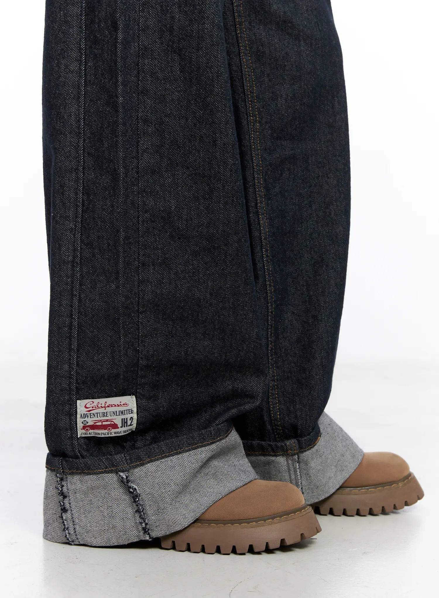 Oakley Cuffed Wide-Leg Jeans CS511 in dark blue, close-up detail of rolled cuff hem, contrast grey inner fabric exposed, California Adventure Unlimited woven patch label, raw edge visible.