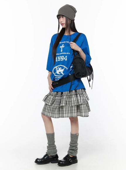 Plaid Layered Midi Skirt CU519 in black, three-quarter front shot on model with black crossbody bag and grey beanie, tiered ruffle plaid skirt styled with oversized blue tee and leg warmers.